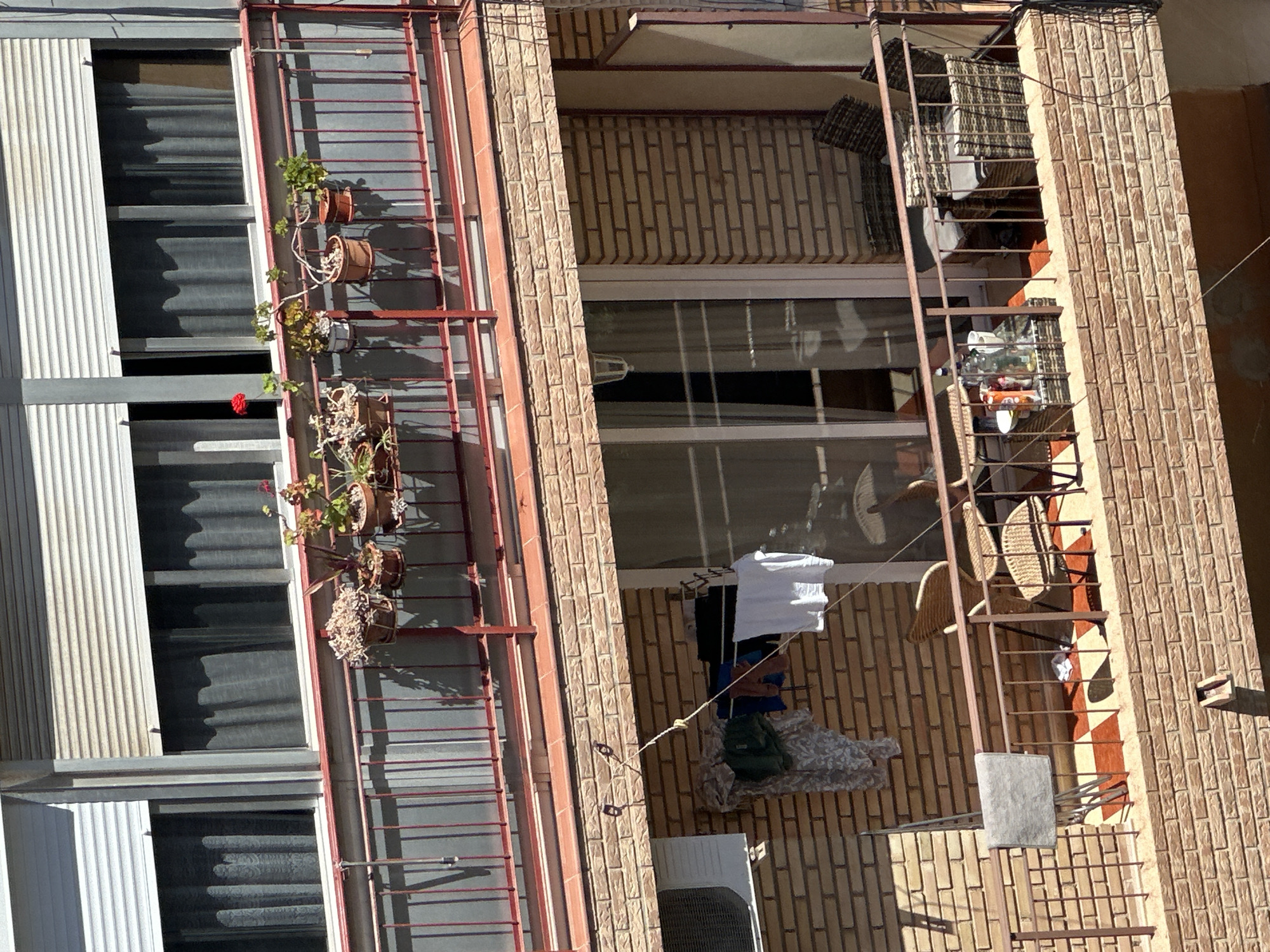 This is a medium-angle, outdoor, eye-level shot of a two-story apartment building exterior. The upper balcony features a red metal railing and several potted plants. The windows above are covered with white roll-down shutters and sheer gray curtains. The lower balcony has a similar red metal railing and is furnished with two wicker chairs, a small table with drinks and snacks, and a wicker couch. There is laundry hanging on a clothesline attached to the brick wall of the building. An air conditioning unit is mounted to the left of the laundry. The building is constructed with light brown brick.