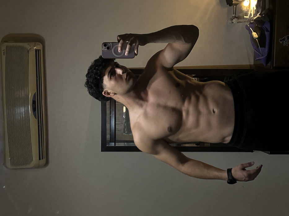 A shirtless man with curly hair takes a selfie in a dimly lit room. His muscular physique, including defined abs, is visible. He's wearing dark pants. A white air conditioning unit is mounted on the wall to his left. A dark-framed object, possibly a mirror or artwork, sits behind him. The lighting suggests a nighttime setting. The man's gaze is focused on the phone in his hand.