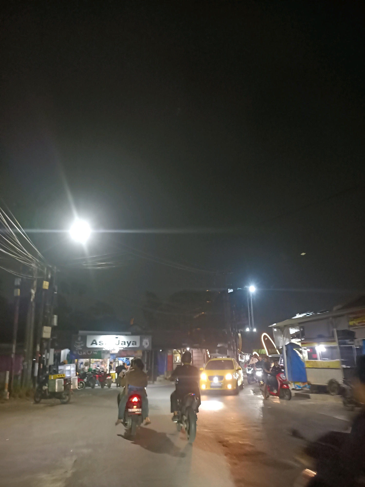 The image captures a nighttime scene on a street in Telagaasih, Indonesia, with a bright street lamp illuminating the road and surrounding structures. Several motorcycles and a yellow car are present, suggesting moderate traffic. On the left side of the street, a storefront with the sign "Asi Jaya" is visible, along with parked motorcycles. The road surface appears wet, possibly from recent rain, reflecting the artificial lights. The sky is dark, indicating nighttime. Two individuals are riding a motorcycle in the foreground, facing away from the camera. Another motorcycle is to their left, and a yellow car is proceeding down the road to the right. The overall impression is a typical urban street scene at night in Indonesia.