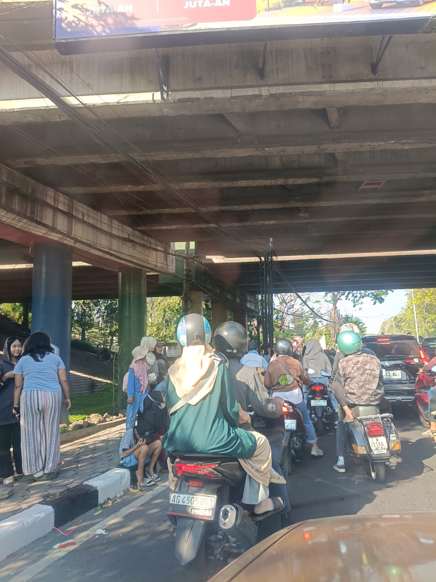 This is a medium shot, vertical, eye-level, outdoor photograph taken in Kembangan, Indonesia. The scene depicts a traffic jam under an elevated road or bridge structure. A convoy of motorcycles and a car are stopped on the asphalt road. People are seated on the motorcycles, some wearing helmets. On the left side of the road, several individuals are standing on the sidewalk. The background shows trees and foliage, indicating an urban environment. The lighting suggests it is daytime, with sunlight filtering through the overhead structure. The overall impression is one of congestion and urban life.