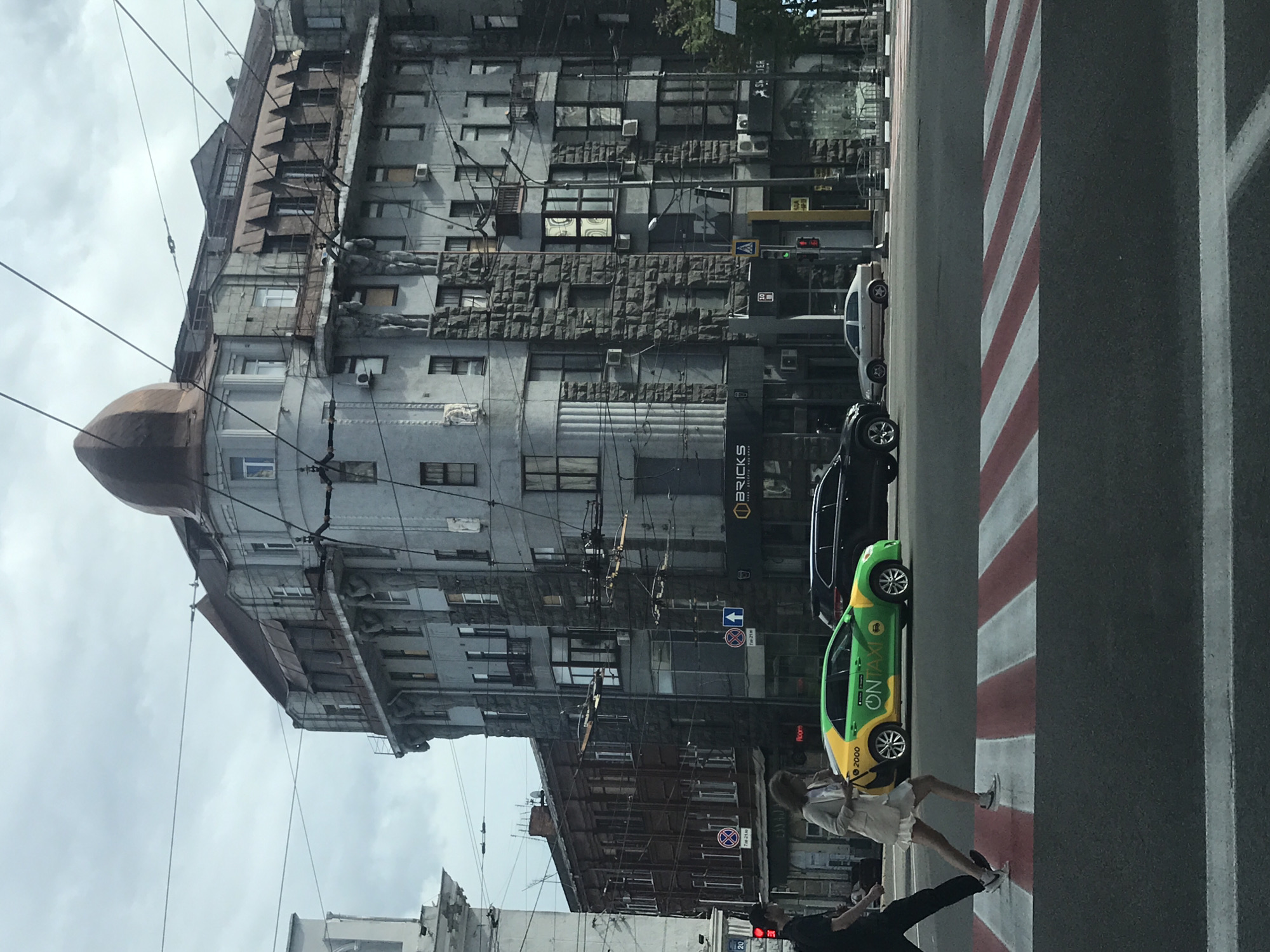 The image depicts a street scene in Kharkiv, Ukraine. A tall, Soviet-era building with a distinctive dome-shaped feature on its corner dominates the left side of the frame. The building's facade is weathered and adorned with numerous windows and air conditioning units. A green and yellow "ONTAXI" branded car is parked along the curb, partially obscuring a "BRICKS" sign on the building. Pedestrians are crossing the street at a crosswalk marked with red and white stripes. Overhead power lines crisscross the view. The sky is overcast.
