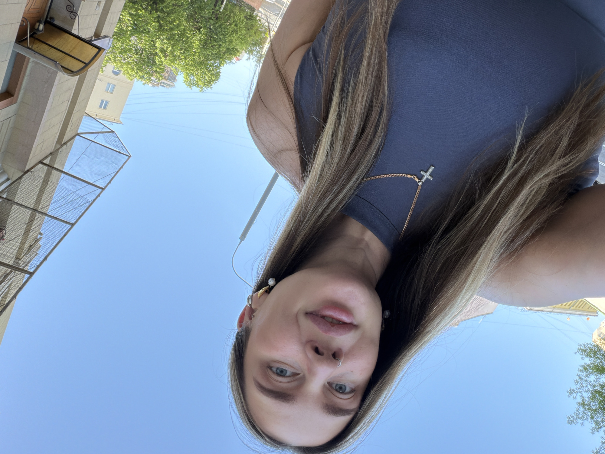 A person with long blonde hair is facing downwards, likely taking a selfie. They are wearing a dark gray top and a gold necklace with a cross pendant. The background features a clear blue sky and parts of a building with an awning and metal grating, as well as a green tree. The location appears to be in an urban area. Moscow, Russia is not discernible from the available information.