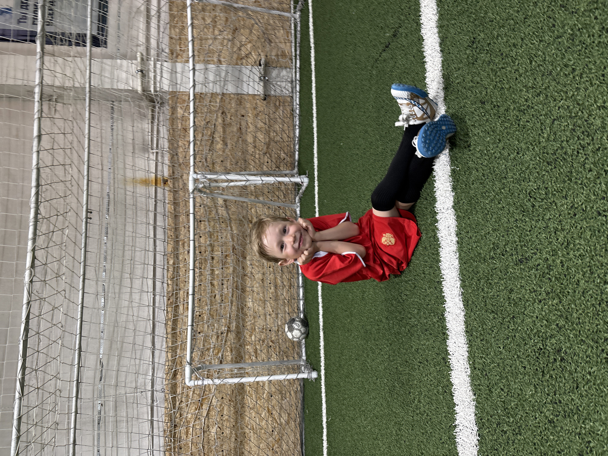 A young child in a red soccer uniform with black leggings and blue cleats lies on a green artificial turf soccer field next to a soccer goal. The child is smiling and has their hands on their cheeks. A soccer ball rests on the edge of the goal. The location is in or near Chaikovsky, Russia, judging by the text “ТЫ ЗДЕСЬ” in Cyrillic visible in the top left corner. The backdrop behind the goal consists of a wall or building structure with light-colored panels and a metal netting system. A white sideline runs along the right edge of the image.