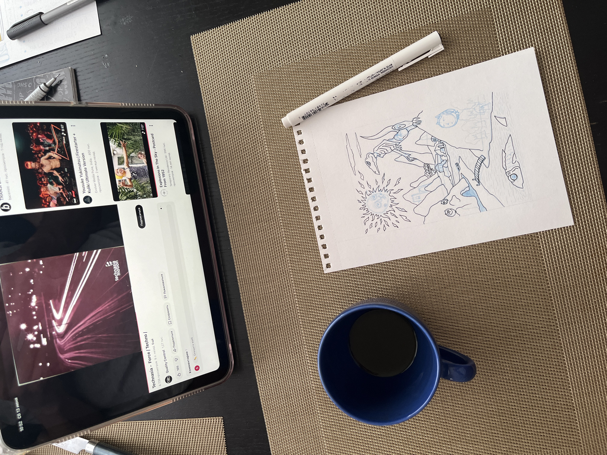 The image depicts a close-up overhead view of a dark brown textured placemat on a table. On the placemat, there is a blue coffee mug filled with a dark liquid, and a sheet of white paper with a pencil sketch. The sketch features a landscape with mountains, a body of water with a fish, a bridge, and stylized figures including what appears to be a dinosaur-like creature and a sun. To the right of the paper is a white pen.
In the background, partially visible, is a tablet device displaying a YouTube interface. The tablet shows two video thumbnails: one with the title "Technasia - Force | Techno |" and another with "Yousuke Yukimatsu | Firestarter | Kuliki Ultimate Version".
The timestamp on the tablet reads "10:52 ОБ 13 ИЮНЯ". "ОБ" likely refers to "обновления" (updates), and "13 ИЮНЯ" means "June 13th". There is no visual information in the image that allows for the identification of the city of Krasnoyarsk, Russia.