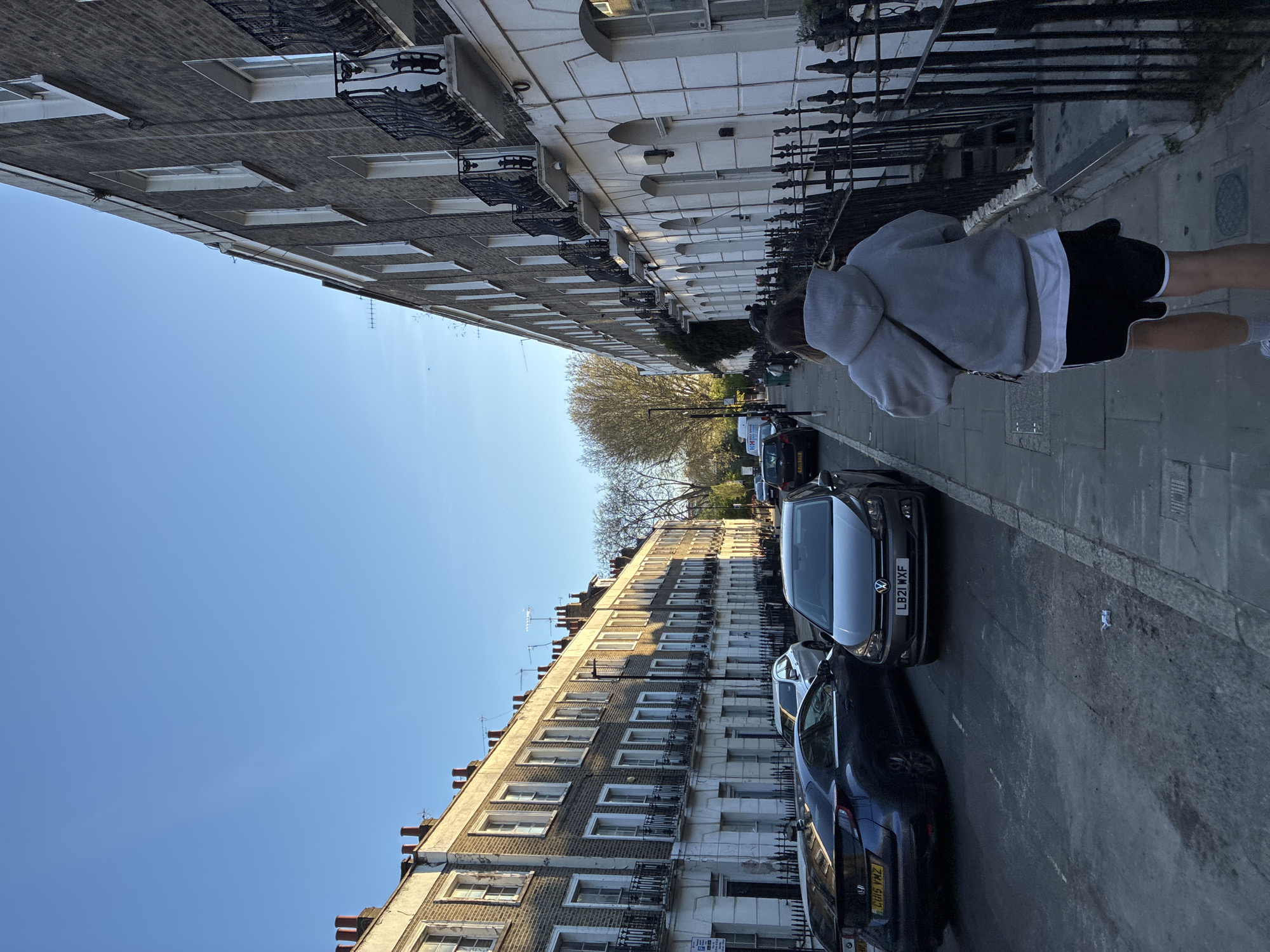 A street scene in London, United Kingdom, shows rows of terraced houses lining both sides of the street. Parked cars are visible along the right side of the road. A person is walking on the sidewalk, wearing a grey hooded sweatshirt and black shorts. The sky is clear and blue.