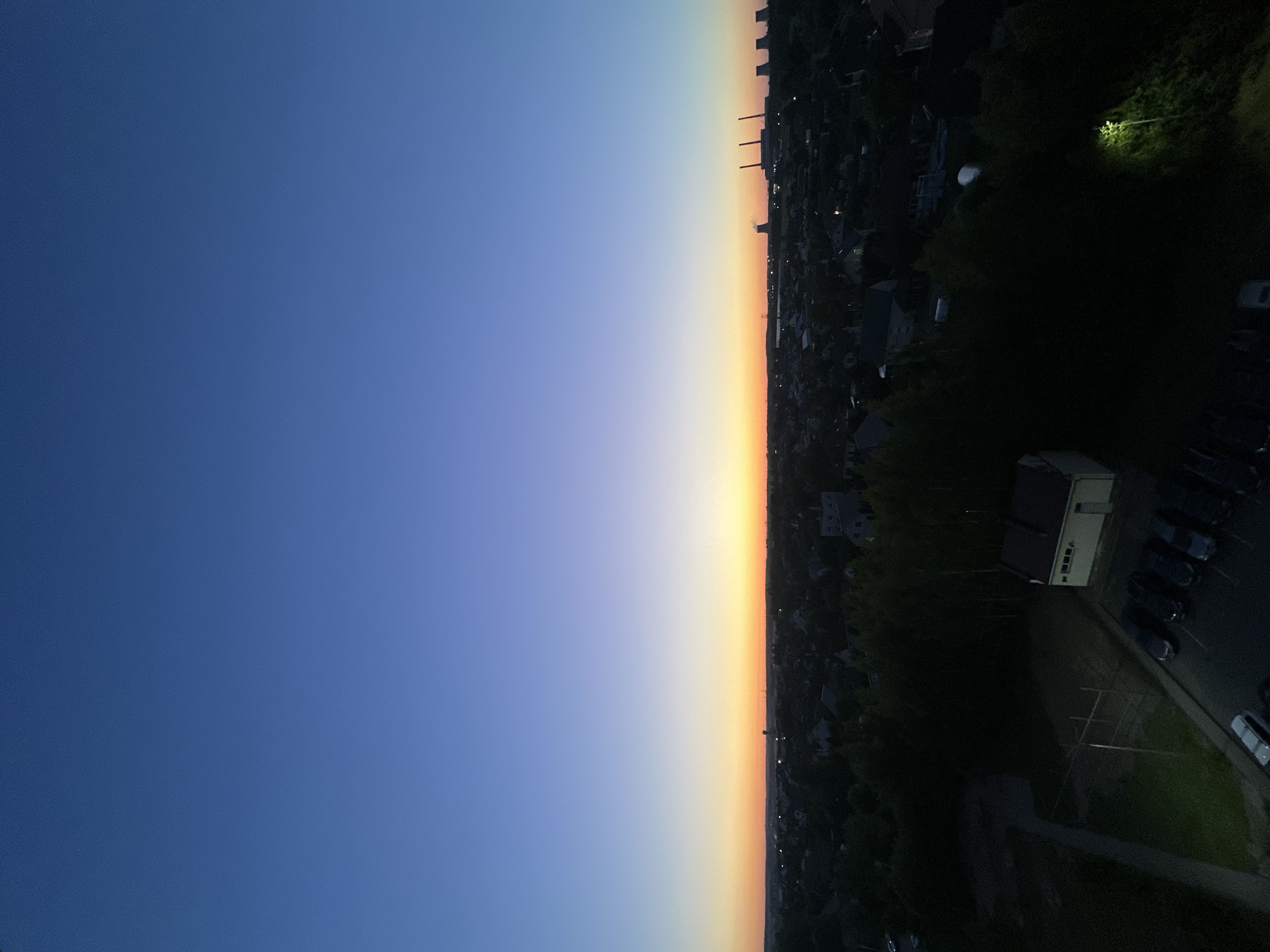 This is a high-angle, wide shot of a suburban landscape at dusk, likely in the vicinity of Шчомысліцкі сельскі Савет, Belarus. The sky displays a gradient of deep blue at the top, transitioning to lighter blues and then to a vibrant orange and yellow band near the horizon, indicating either sunrise or sunset.
Below the sky, a dense array of trees forms a dark canopy over a residential area. Scattered houses with visible rooftops are interspersed within the foliage. In the middle ground, several industrial structures with tall chimneys are silhouetted against the colorful sky on the right side of the frame.
In the foreground, on the left side, a paved area shows a row of parked cars, mostly dark in color. A small, light-colored building with a dark roof is situated in the lower center of the image, with a light emanating from a streetlamp nearby, illuminating some of the surrounding vegetation. The overall impression is a quiet, transitional period of day in a developed, yet green, urban fringe.