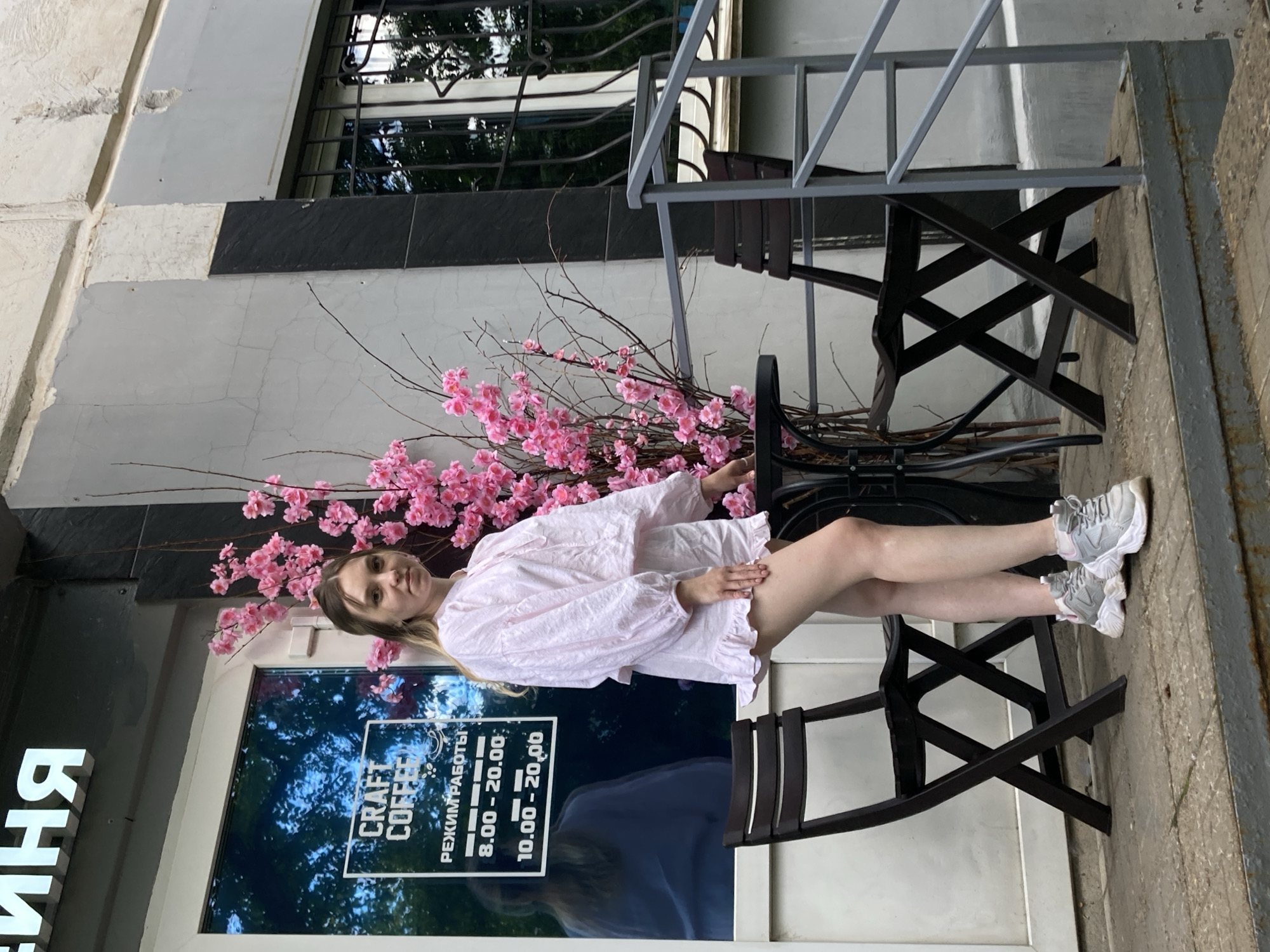 A young woman with blonde hair sits on a black chair outside a cafe, her legs crossed. She is wearing a loose, light pink dress and grey sneakers. Behind her, a display of artificial pink cherry blossoms arches over her. She is looking directly at the camera with a neutral expression.
The cafe has a large window with "CRAFT COFFEE" written on it in white lettering, along with operating hours. The surrounding environment appears to be urban, with a building facade and a metal railing visible. The lighting suggests it is daytime, possibly late afternoon, given the soft shadows. The weather seems pleasant enough for outdoor seating.
Visible text includes "CRAFT COFFEE" and "РЕЖИМ РАБОТЫ" followed by times "8.00 - 20.00" and "10.00 - 20.00". There are also large white letters on the left side of the image, partially visible, which appear to be part of a sign for the establishment. The overall scene depicts a casual moment at an urban cafe.