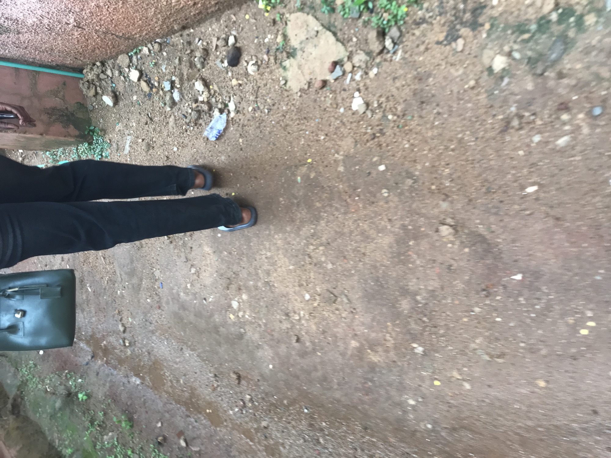 A person is walking away from the camera down a narrow, unpaved path in Jos, Nigeria. The ground is a mix of dirt, gravel, and scattered debris, with some puddles indicating recent rain. To the right of the path, a rough, reddish-brown wall is visible, with a vertical green pipe running along it. The person is wearing dark trousers and dark sandals. They are carrying a dark green handbag in their left hand. The overall environment appears to be a less developed or residential area of the city, possibly an alleyway or side street.