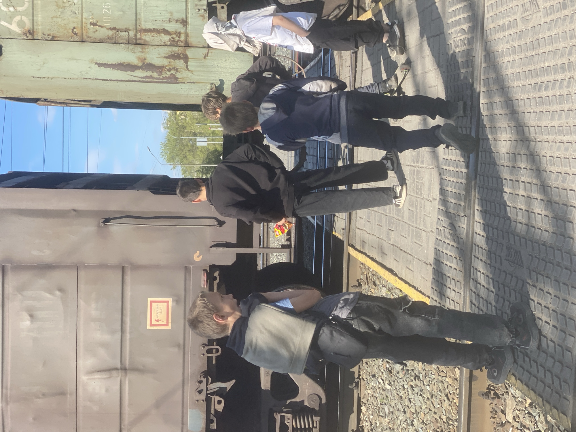 The image depicts a group of four people standing next to a train car on a train platform. The people appear to be male. The train is gray. The platform is made of concrete with a textured surface. The train is located in Novokuznetsk, Russia.