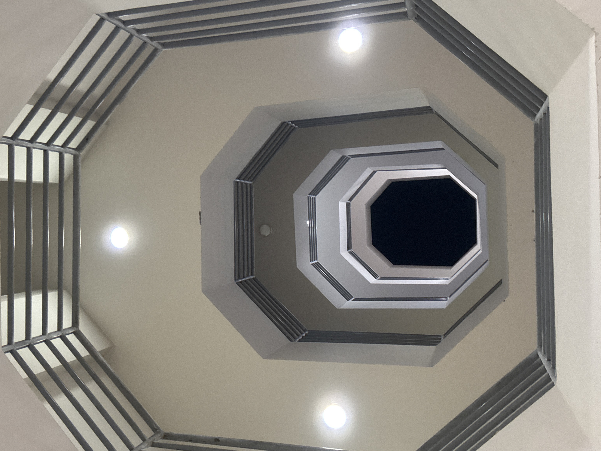 The image depicts an upward view into a stairwell with an octagonal shape. The stairwell is illuminated by three evenly spaced circular lights, each emitting a bright glow. The walls are painted in a light beige or cream color. The architecture consists of a series of receding octagonal levels, creating a tunnel-like effect leading to a dark opening at the top. Each level features a trim composed of parallel metal bars, adding a linear design element to the structure. The overall effect is a perspective drawing the eye upwards towards the central opening.