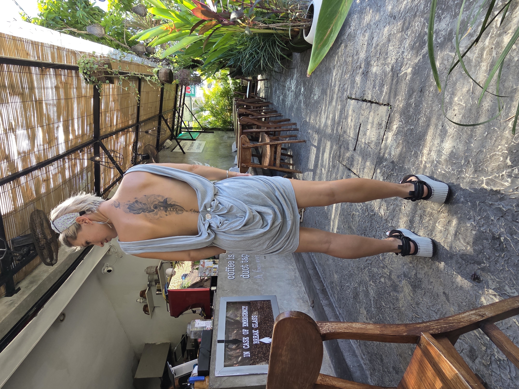 Here is a detailed description of the image:
**Scene Overview:**
The photo appears to be taken at an outdoor café or coffee shop, possibly in a tropical location, as indicated by the lush greenery. The perspective suggests the photo was taken at ground level or slightly below, looking upwards.
**People:**
- There is one person visible - a Caucasian woman in a light blue dress. She has blonde hair and is wearing a headband. She has a tattoo on her back.
**Objects:**
- **Furniture:** Wooden chairs and tables are present, typical of a café setting.
- **Plants:** Various types of plants and foliage are seen in the background and overhead. Some plants are in hanging baskets.
- **Café Elements:** A coffee machine and other equipment are visible. There's a sign with the text "Coffee is duct tape. It fixes every..." and another stating "In case of emergency break glass."
- **Shoes:** The woman is wearing sandals with a thick, light-colored sole and black straps.
- **Structure:** The café area seems to have a bamboo or woven roof, providing shade.
- **Accessories:** There appears to be a headband and earrings.
**Environment:**
- **Location:** Based on the style of the café, tropical plants, and local furniture, this could be in a tourist area. It is in North Kuta, Indonesia.
- **Lighting:** The scene has bright, natural lighting, with some areas in shade.
- **Surfaces:** The ground appears to be paved with a textured, grey-colored material, possibly stone or concrete.
**Actions/Interactions:**
The woman seems to be standing or posing near the coffee bar.
**Additional notes:**
- The overall impression is a relaxed, outdoor café setting in a warm climate.