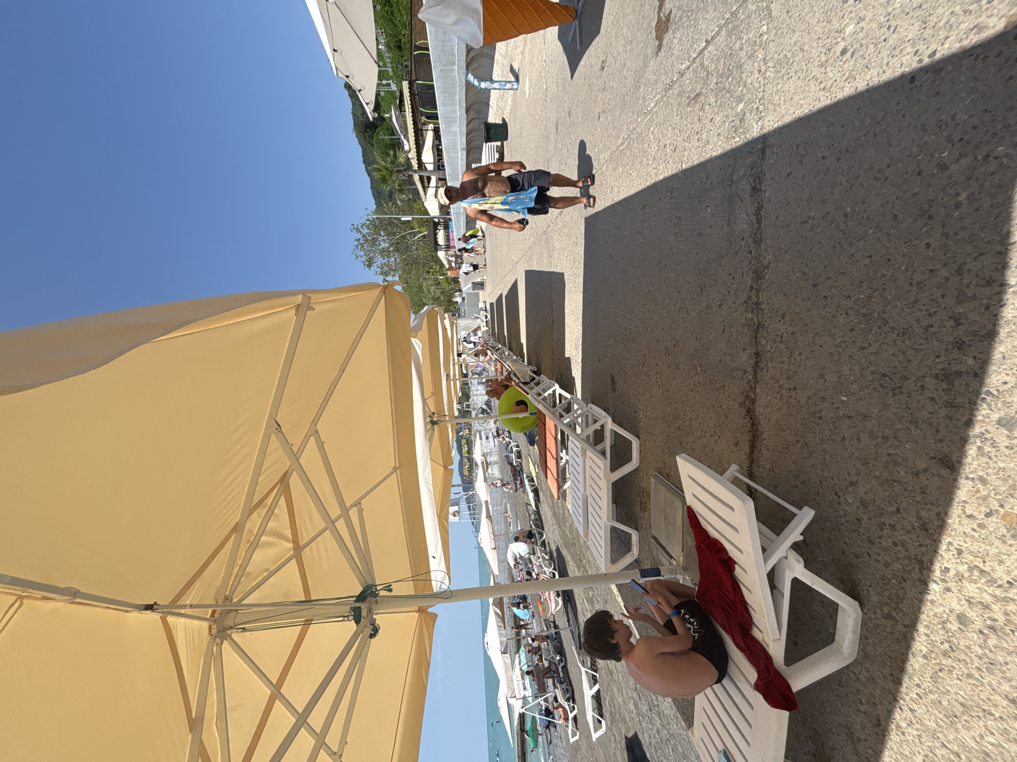 A sunny day on a pebble beach in Sochi, Russia, with a promenade lined with lounge chairs and large beach umbrellas. In the foreground, a young boy sits on a white lounge chair, engrossed in his phone. A red towel is draped over the chair. Further down the promenade, several other beachgoers recline on lounge chairs under umbrellas, some facing the sea. To the right, a man with a towel slung over his shoulder walks along the promenade. In the background, the Black Sea stretches out under a clear blue sky, with a few boats visible on the water. Hills covered in greenery rise behind the beach. A large decorative ice cream cone statue is visible on the far right.