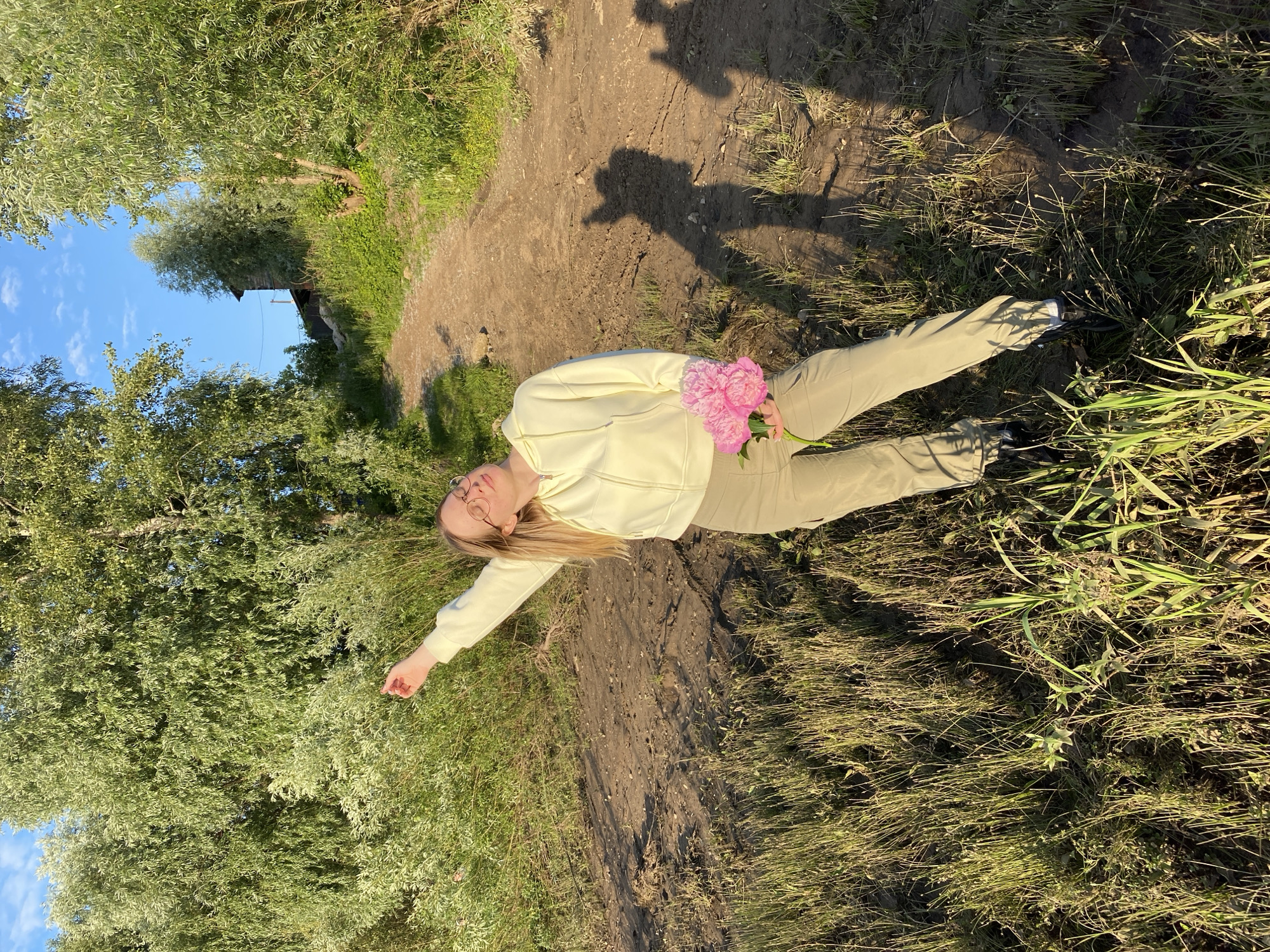 A young woman with blonde hair and glasses stands on a dirt path in a grassy area near trees, holding a bouquet of pink peonies. She is wearing a light yellow hooded jacket and light brown cargo pants. Her left arm is raised and her eyes are closed. The scene appears to be outdoors, with a dirt path winding uphill and lush green vegetation. Shadows are cast on the ground, suggesting it is late afternoon or early evening. The location is identified as Chaikovsky, Russia.