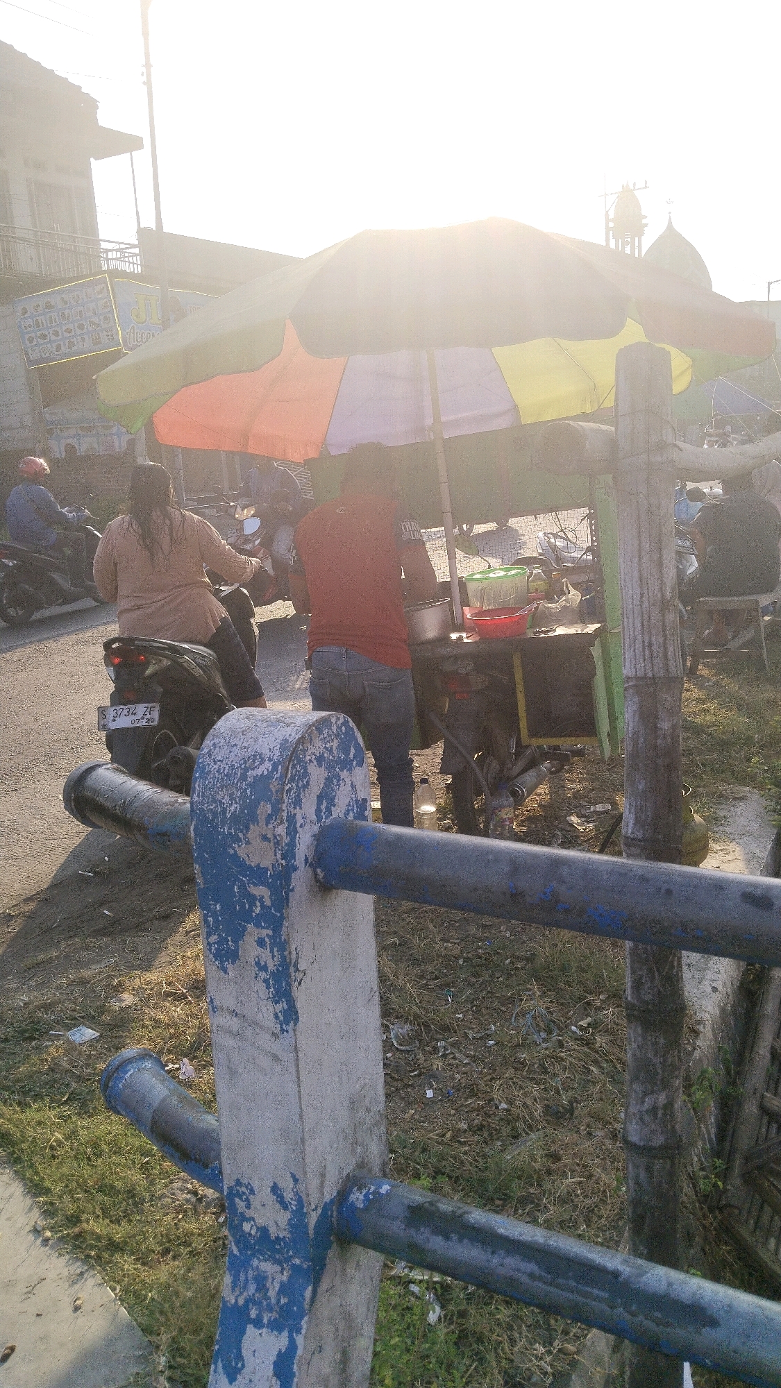 The image depicts a street scene in Kedawong, Indonesia, under bright daylight conditions, suggesting it is daytime. The foreground features a concrete pillar with weathered blue paint, supporting horizontal blue metal bars, part of a railing or barrier. Behind this, a black motorcycle with license plate "S 3734 ZF" is parked. A person in a light brown top and dark pants is seated on the motorcycle, facing away from the camera. To the right of the motorcycle, a street food vendor operates from a cart. A person wearing a red t-shirt and blue jeans stands behind the cart, appearing to prepare or serve food. The cart is covered by a colorful umbrella with sections of orange, yellow, and green. Visible on the cart are containers, possibly for food or drinks, including a green-lidded container and a red bowl. A bottle of water is on the ground near the cart. In the background, a building with a balcony is visible, and further in the distance, a mosque with a dome and minaret is present, identifying the location as Kedawong. Other motorcycles and people are also present in the scene, with some figures blurred due to motion or distance. The overall environment appears to be a roadside setting with dry grass and scattered debris.
