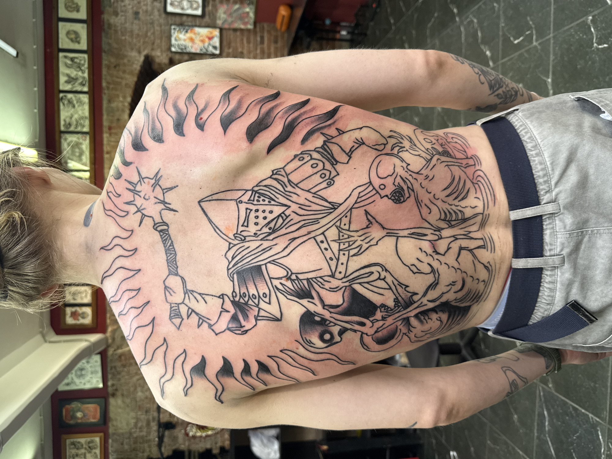 The image shows the back of a person with a large black ink tattoo covering their upper back and shoulders. The tattoo depicts a figure in armor, possibly a knight, wielding a spiked mace. Surrounding the armored figure are several skeletal or zombie-like creatures. The tattoo design includes dynamic lines suggesting flames or energy emanating from behind the armored figure. The person is wearing light gray pants with a dark blue belt. The background appears to be an interior space with brick walls and framed artwork or posters hanging on the walls. The flooring is dark and reflective. This scene is likely in a tattoo parlor in Moscow, Russia.