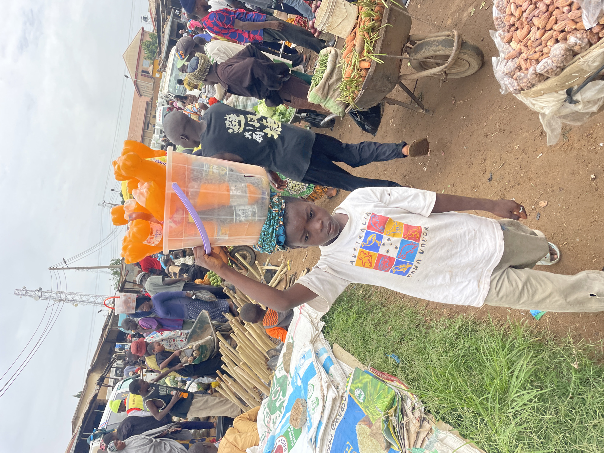 A young boy walks through a busy outdoor market in Gura Top, Nigeria, carrying a large plastic bucket filled with orange beverages and cans on his head. He wears a white t-shirt with a colorful "Australia Down Under" graphic and light-colored pants. To his right, a man in a black t-shirt with white text walks with another bucket on his head. In the background, a wheelbarrow is filled with carrots and green beans. Several other people, dressed in various casual attire, are seen browsing and interacting within the crowded marketplace, which features stalls and a white van. The ground is dusty and red, and stacks of white sacks and bamboo poles are visible. The sky is overcast.