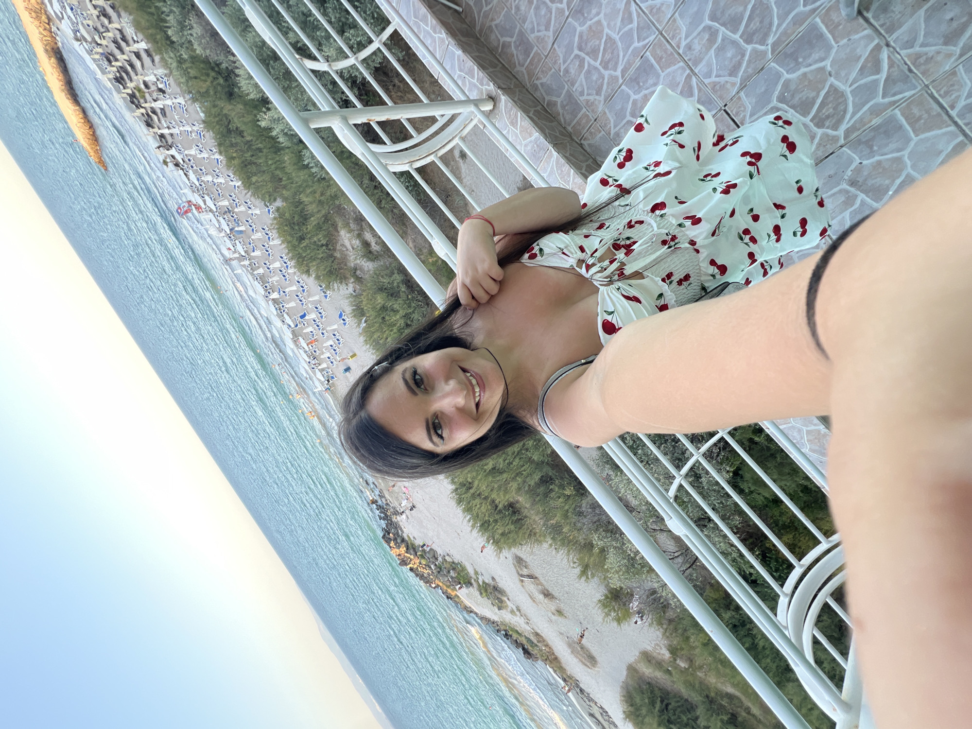 A woman with long dark hair is taking a selfie on a balcony overlooking a beach and the sea in Pomorie, Bulgaria. She is wearing a white dress with a cherry print. The beach is lined with white lounge chairs and umbrellas, and the sea is calm and blue. The sky is clear and a pale blue. The balcony has a white railing and a tiled floor. There are some people on the beach, but the focus is on the woman.