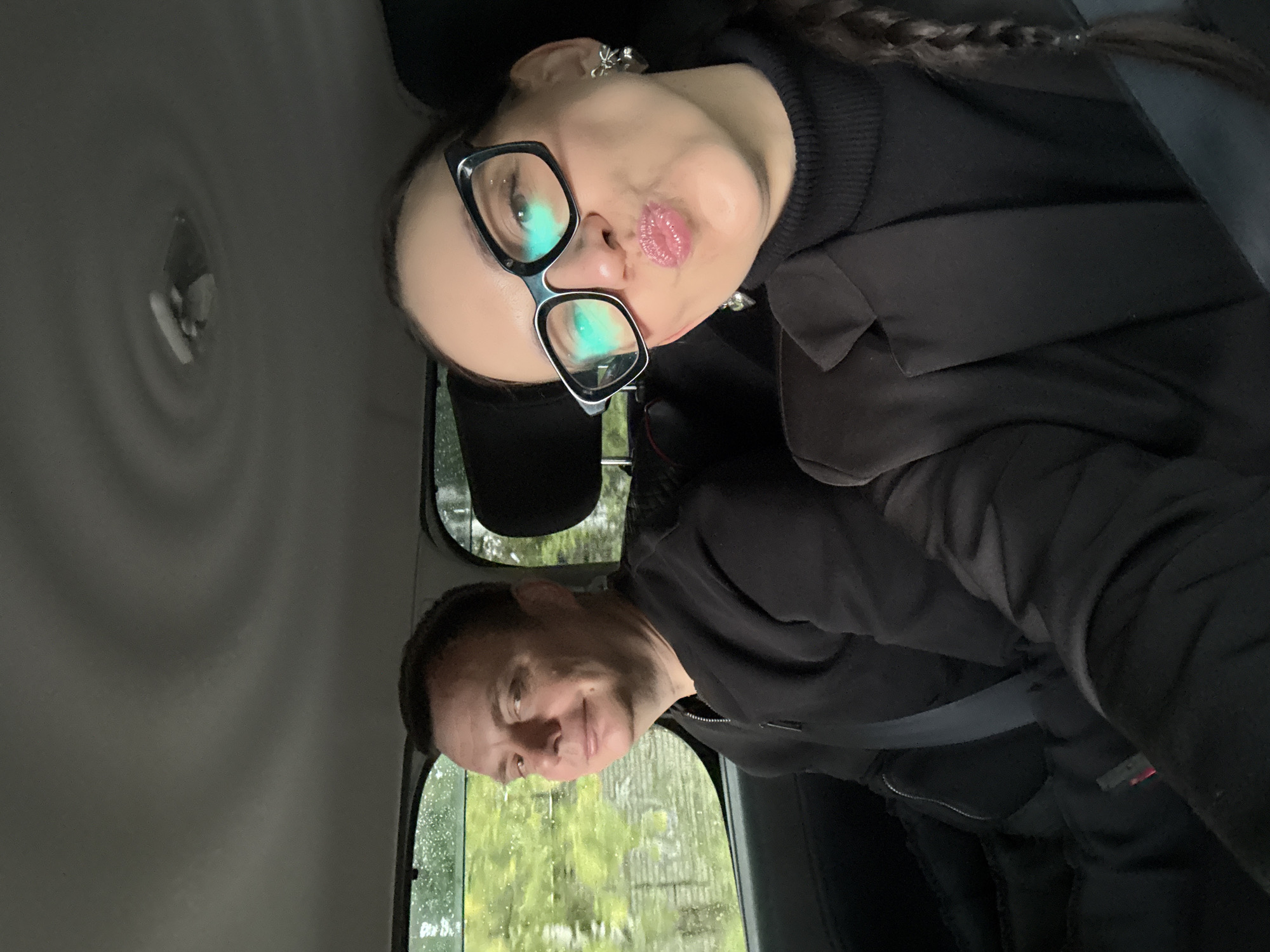 Two individuals, a man and a woman, are seated inside a vehicle. The man, positioned on the left, is looking towards the camera with a slight smile. He is wearing a black zip-up jacket and a seatbelt. The woman, on the right, is leaning towards the camera, making a kissing face, and wearing glasses, a black turtleneck, a black blazer, and heart-shaped earrings. Her hair is styled in a braid. Both are wearing seatbelts. The interior of the vehicle, including the ceiling and seats, is visible. Outside the car windows, a blurred green landscape with trees is apparent, suggesting it is daytime and possibly raining due to the visible droplets on the window. The overall setting appears to be a street in Chaikovsky, Russia, given the context.