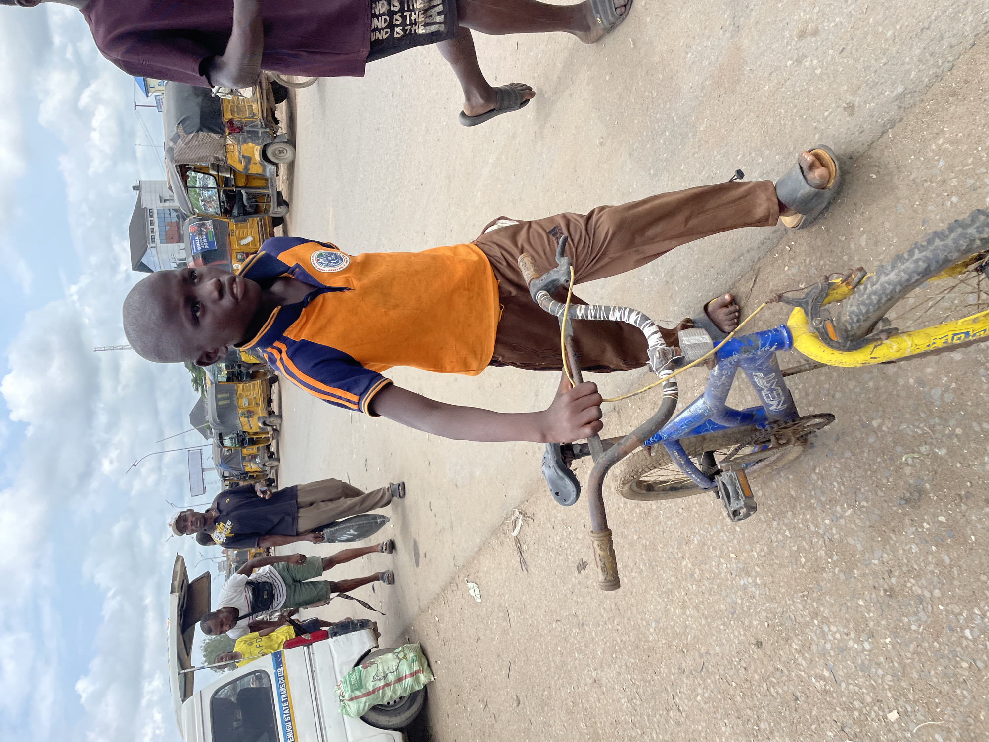 In Onitsha, Nigeria, a young boy with a shaved head, wearing an orange and blue collared shirt and brown pants, walks with a blue and yellow bicycle. He is holding the handlebars of the bicycle, which appears to be well-used and possibly dirty. To his right, another person, partially visible, wears dark shorts with white lettering and dark sandals. In the background, several yellow motorized tricycles, commonly known as 'kekes', are parked or in motion on a paved road. Other individuals are also present in the scene, some near a white van with a covered roof. The environment appears to be a busy urban street with a cloudy sky overhead.