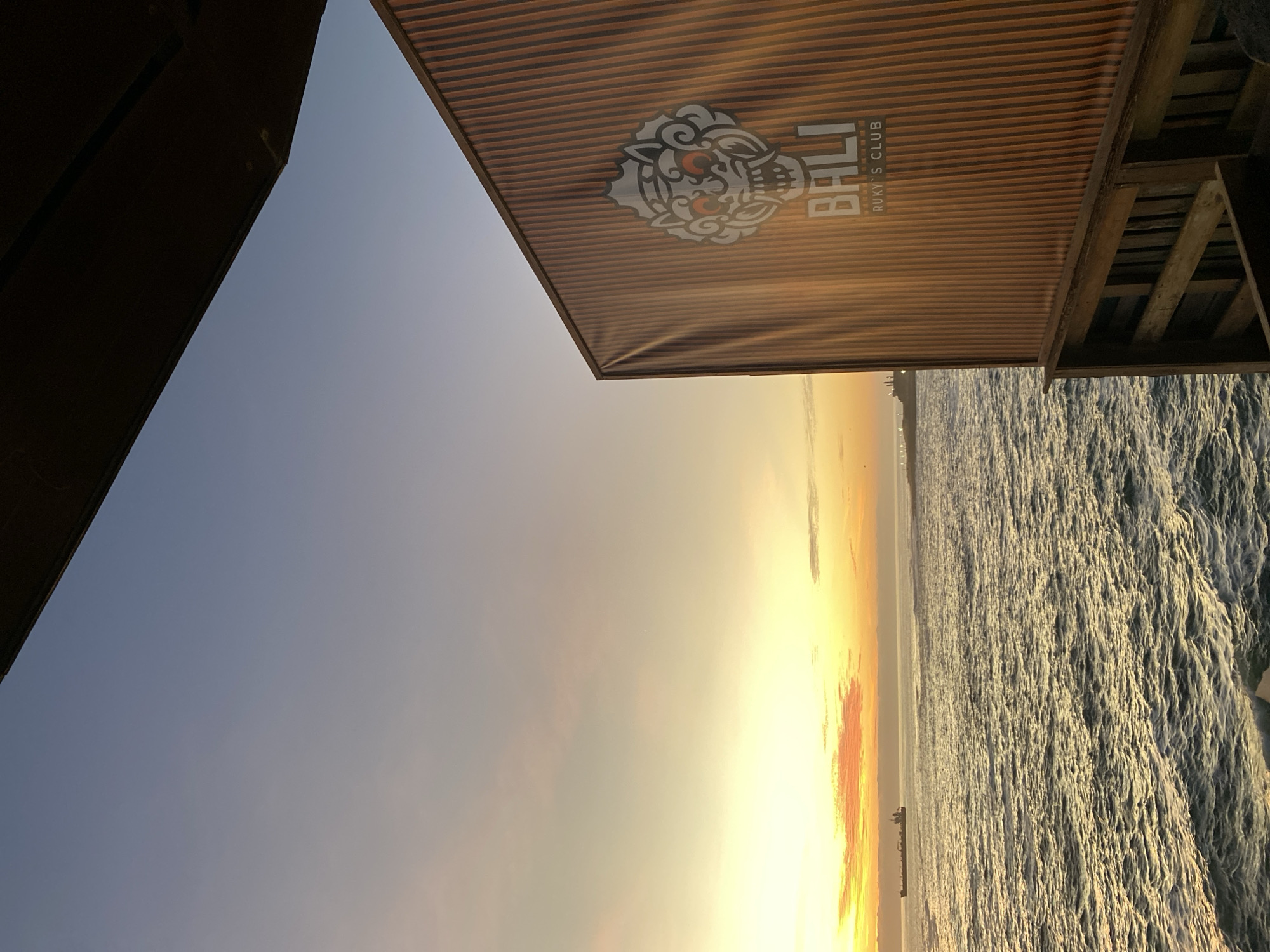 The image shows a sunset over the Pacific Ocean in Arica, Chile. The perspective is from under a wooden structure, with a sign for "BALI Ruky's Club" prominently displayed. The sun is setting on the horizon, creating a bright glow with orange and yellow hues reflecting on the water. The ocean surface is textured with small waves. A ship is visible in the distance. The sky transitions from a lighter, golden color near the horizon to a clear blue at the upper left.