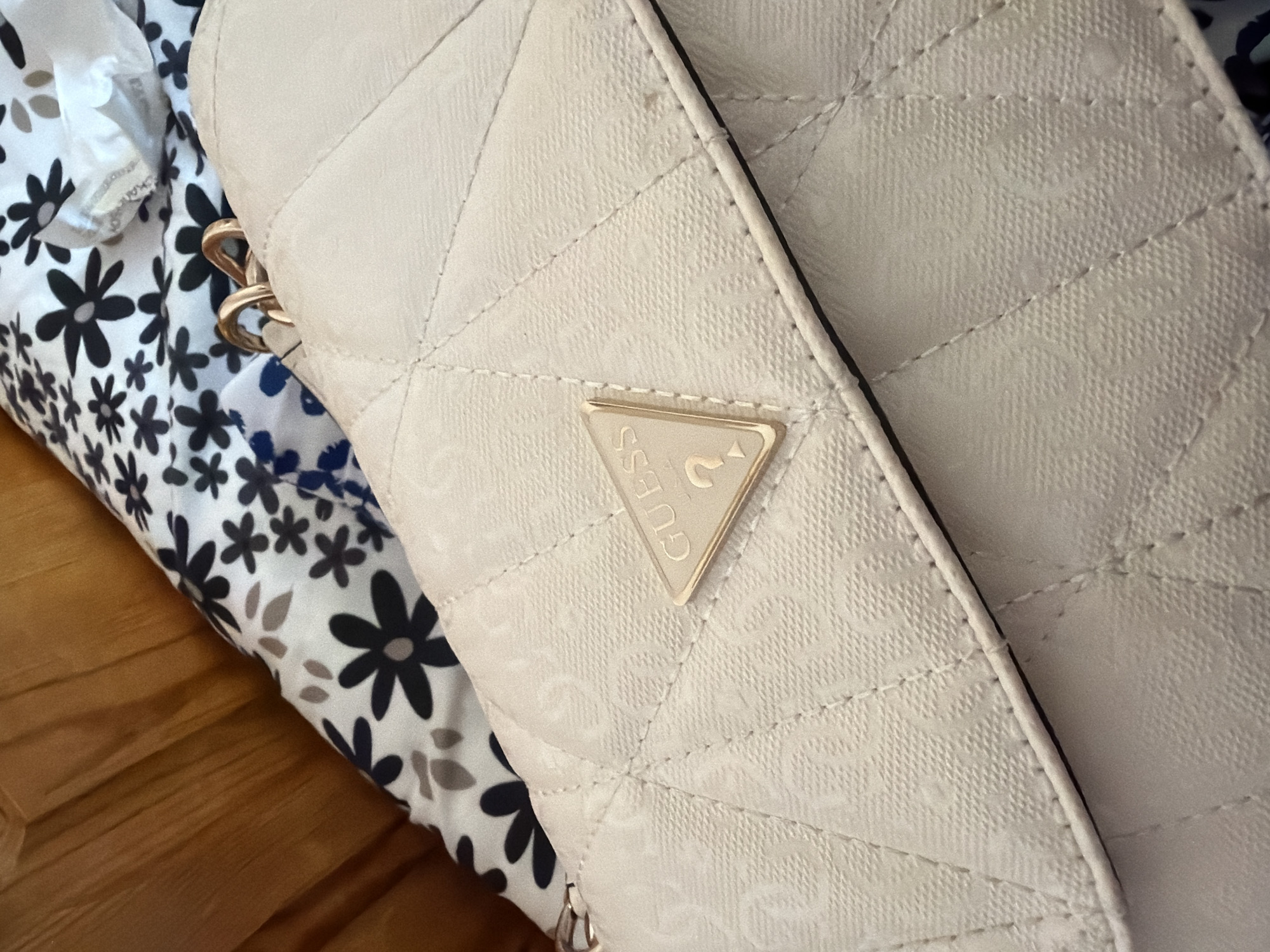 A close-up shot captures a light-colored, quilted bag, likely a purse or wallet, resting on a patterned fabric and a wooden surface. The bag is cream or beige, featuring a diamond-shaped quilted pattern. A subtle, textured design, possibly a repeating logo, is visible across its surface. A prominent gold-toned metallic triangular emblem is affixed to the front, clearly displaying the word "GUESS" and the brand's iconic upside-down triangle symbol. Partial gold-colored chain links, suggesting a strap, are visible on the upper left side, and a silver-toned zipper pull can be seen at the bottom. The bag lies against a white fabric background adorned with black and some gray/brown floral patterns. A wooden surface, possibly a floor or table, occupies the lower left corner of the frame. No individuals are present, and no specific activity is taking place.