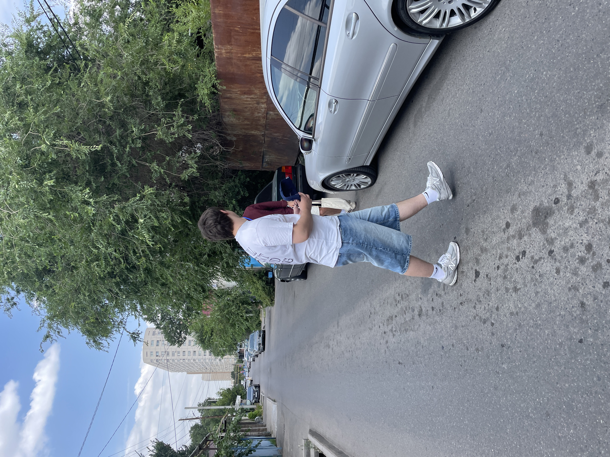 A person stands on an asphalt road, facing away from the camera and towards a silver car. The person is wearing a white t-shirt, denim shorts, and white sneakers. They appear to be holding a mobile phone. The scene is outdoors, with a large tree to the left and a rusty metal structure behind the car. In the background, there are parked cars, more trees, and a tall, light-colored building under a partly cloudy sky. The image seems to have been taken during the day, likely in the summer, given the foliage and attire of the person. The road is a typical urban street, with some discoloration and oil stains visible on the asphalt. The overall impression is a casual, everyday scene in an urban environment.