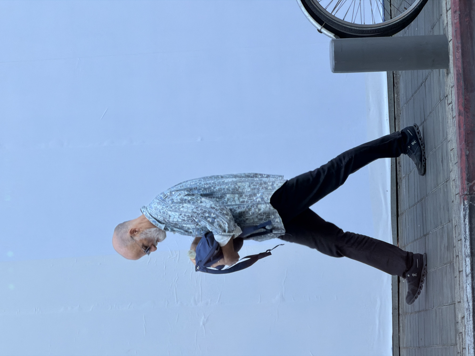 A middle-aged, bald man with a gray beard is seen holding a navy backpack. He is wearing a patterned, light-blue shirt, dark pants, and black shoes. He stands at an angle on a sidewalk made of gray bricks next to a gray, cylindrical bike rack. The background is a white wall. The photograph was taken in Tel-Aviv, Israel.