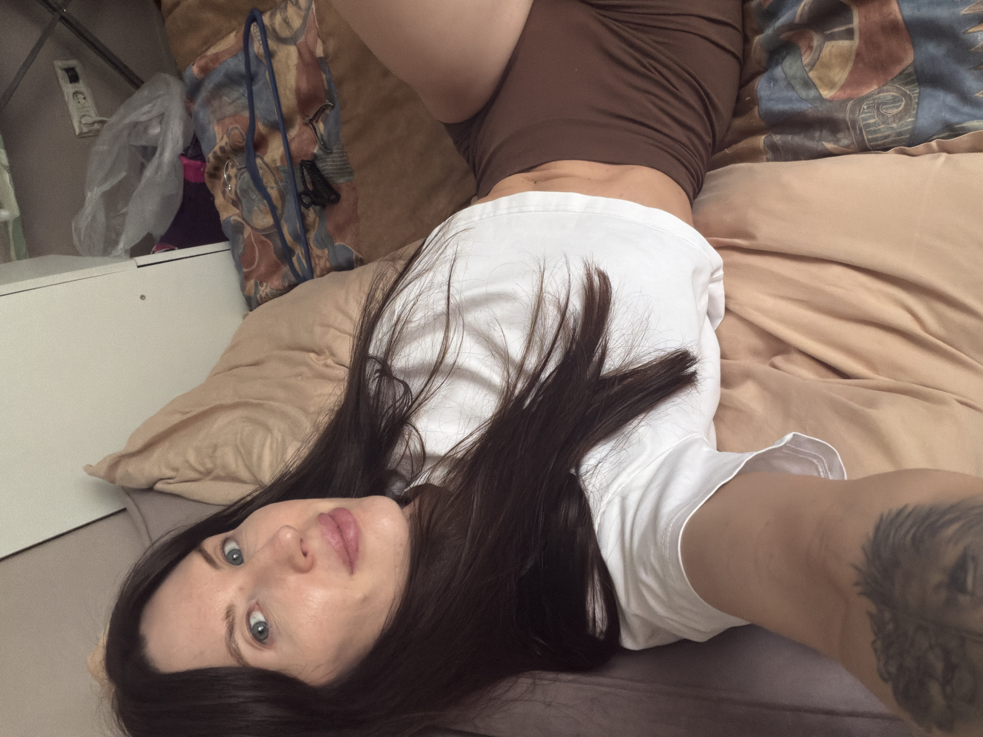 A woman with long, dark hair is lying on a bed with tan-colored sheets and pillows. She is wearing a white t-shirt and brown shorts. A tattoo is visible on her right arm. Background details include a dresser, clothes hangers, and a patterned pillow. There is no information to suggest the image was taken in the city of Perm, Russia.