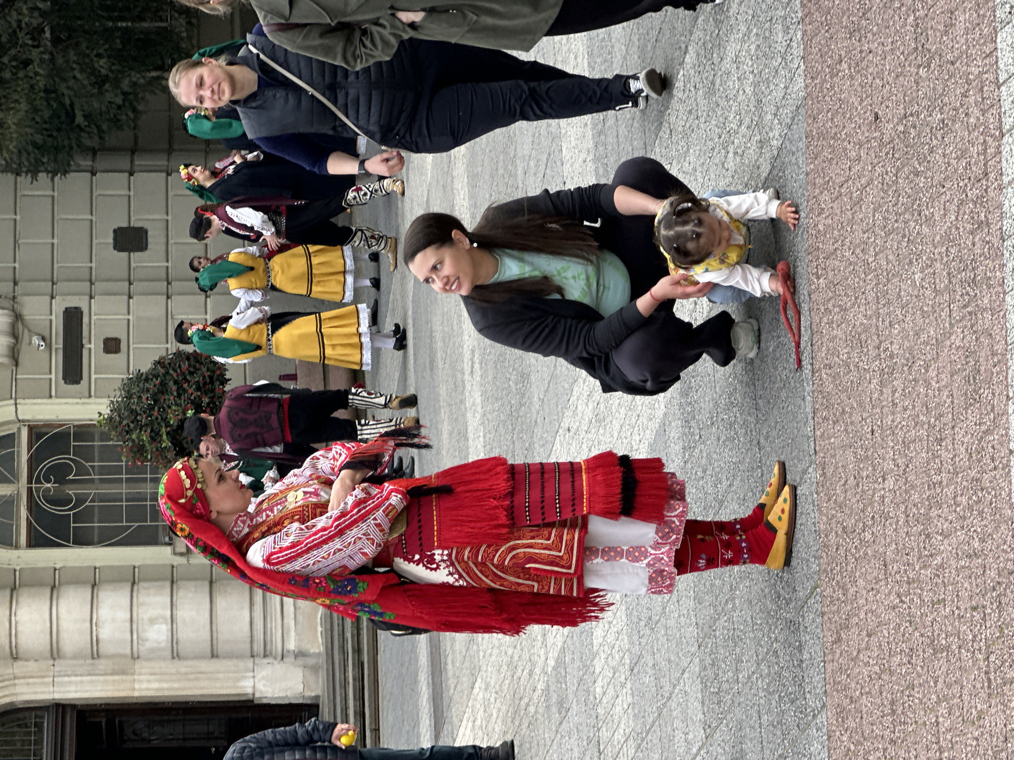 The image shows a scene in what appears to be the city center of Plovdiv, Bulgaria. In the foreground, a woman dressed in traditional Bulgarian folk attire with a red and white patterned outfit, a red headscarf, and yellow shoes is standing on a light gray and pink patterned pavement. To her right, a younger woman is kneeling on the pavement, holding a baby in her lap. Behind them, a group of people are standing and dressed in traditional Bulgarian folk costumes. The backdrop includes a building with a decorative window and a small tree or bush near the building. Additional people are also visible in the background. The scene appears to depict some sort of cultural gathering or performance.