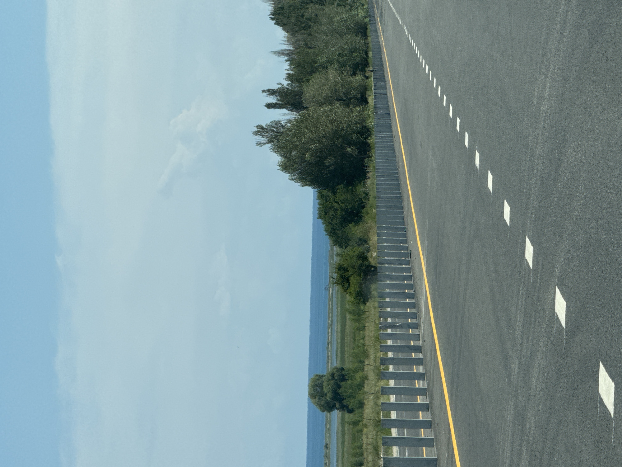 The image depicts a road with a concrete barrier and yellow line on the left side, and a body of water and trees in the background under a cloudy sky. The road has white dashed lines painted on it, indicating lane markings. The scene appears to be located in or near the city of Kara-Talaa, Kyrgyzstan, given the context provided. There are no people or animals visible in the image. The environment is a roadside with natural elements in the distance.