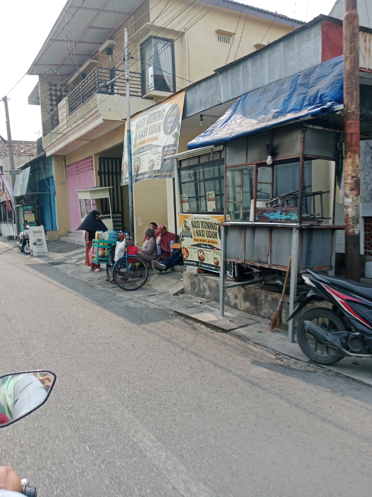 The image captures a street scene in Kediri City, Indonesia, featuring a row of buildings with shops and food stalls. In the foreground, a street with asphalt paving is visible. To the right, a black and red motorcycle is parked. Further down the street, a blue tarp covers a food stall with metal framing and glass windows. A broom leans against the stall. Adjacent to the stall, a banner advertises "Nasi Kuning & Nasi Uduk" (yellow rice and mixed rice). On the sidewalk, several people are seated, and a woman in a black hijab stands near a bicycle laden with food containers. Another food stall, advertising "Bakso Solo", is visible with a motorcycle parked in front of it. The buildings appear to be two stories tall, with balconies on the upper level of one. The sky is overcast.
