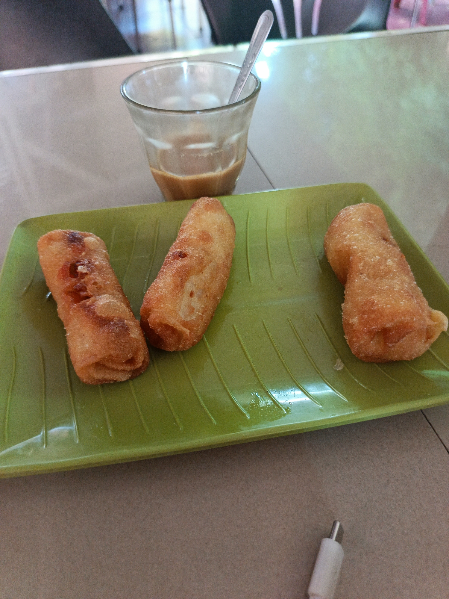 The image depicts a tabletop scene with a light green, leaf-patterned tray holding three fried spring rolls. Behind the tray is a glass with a spoon in it, containing a milky brown liquid, likely coffee or tea. A partial view of a white charging cable is present in the bottom right corner of the frame. The setting appears to be a casual dining environment, possibly a cafe or restaurant in Lhokseumawe, Indonesia, given the regional cuisine.