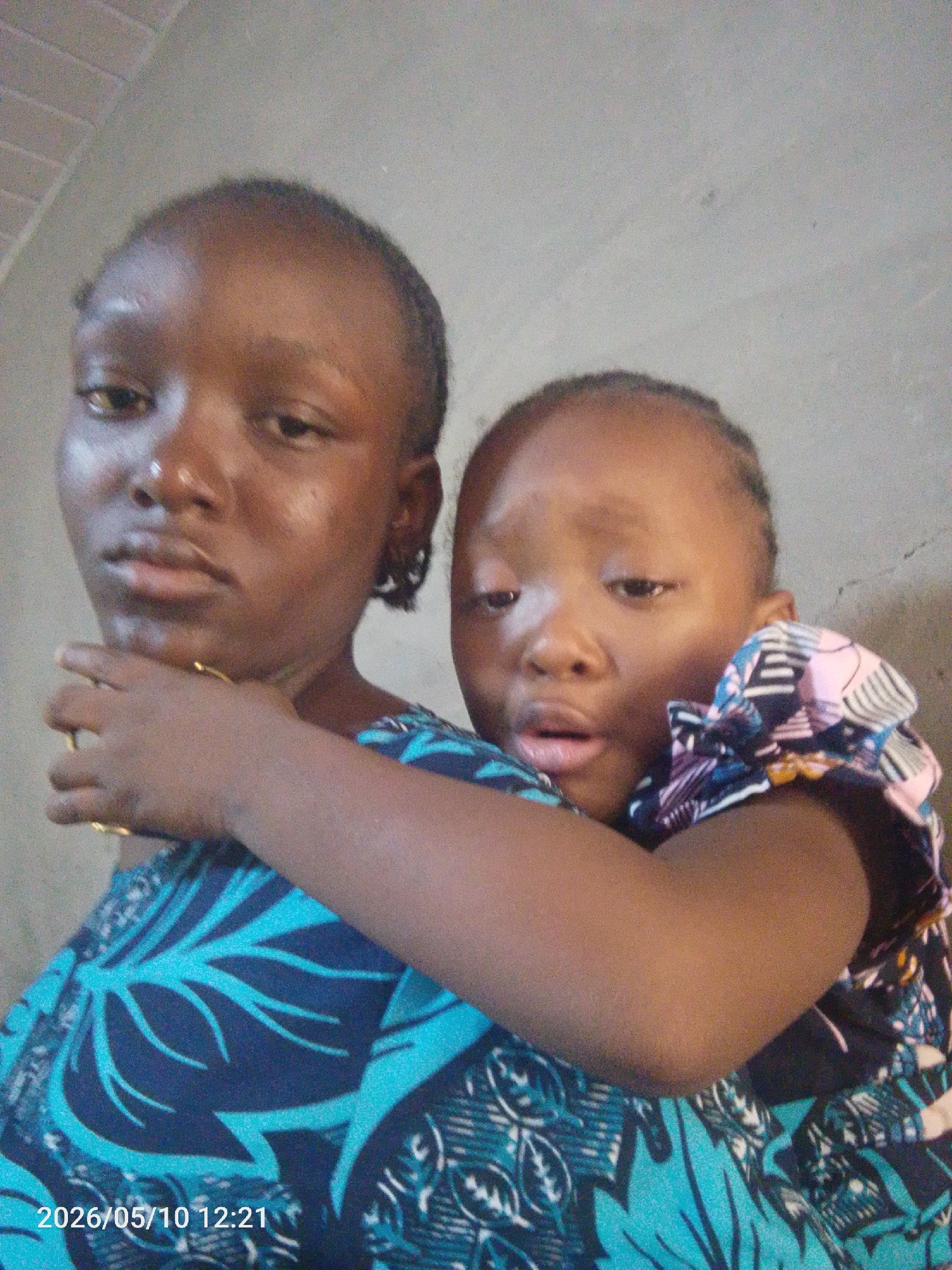 Two dark-skinned females are in a close embrace, with a lighter colored wall as background. The image has a date stamp of 2026/05/10 12:21. The older female is wearing a bright blue patterned dress, and she has a gold bracelet on her left wrist. The younger female is wearing a patterned top with white, black, pink, and blue colors. Their faces show a serious, slightly sad, expression. Location: Lokoja, Nigeria.