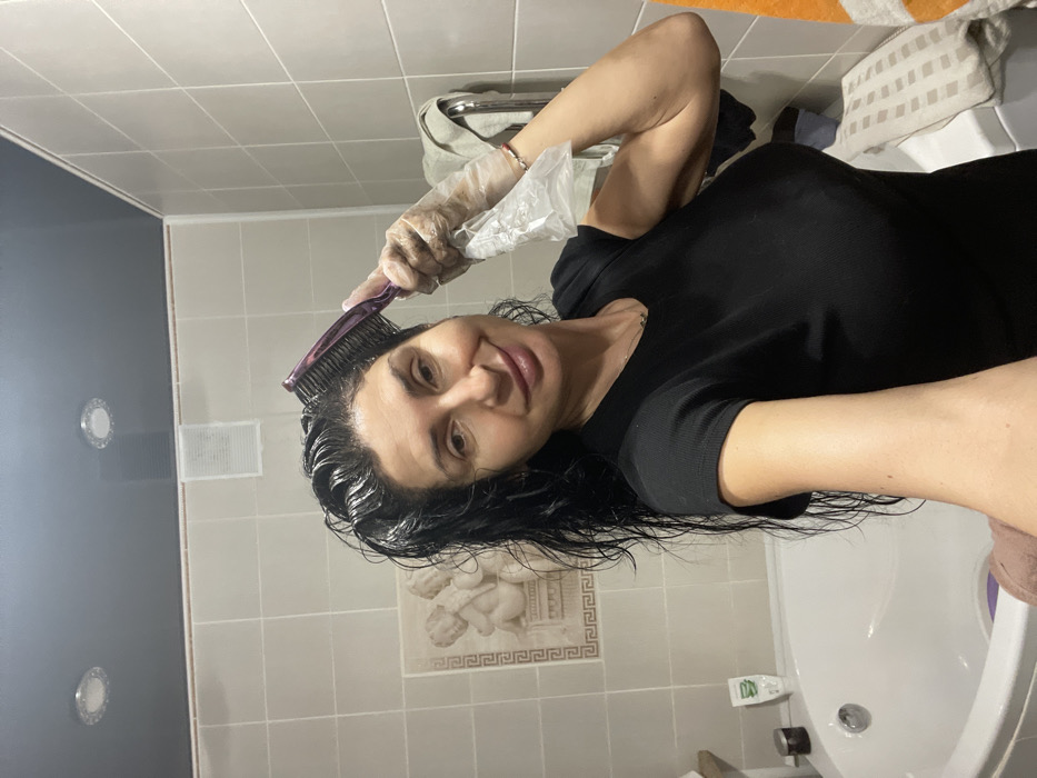 A woman in a black top applies hair dye with a purple comb while wearing a plastic glove. She is in a tiled bathroom with a white tub, and her wet hair is slicked back. Bathroom fixtures include a white vent and recessed lighting on the left side of the image. A towel and bag rest above the woman, near the upper edge. The woman is smiling and facing the camera.