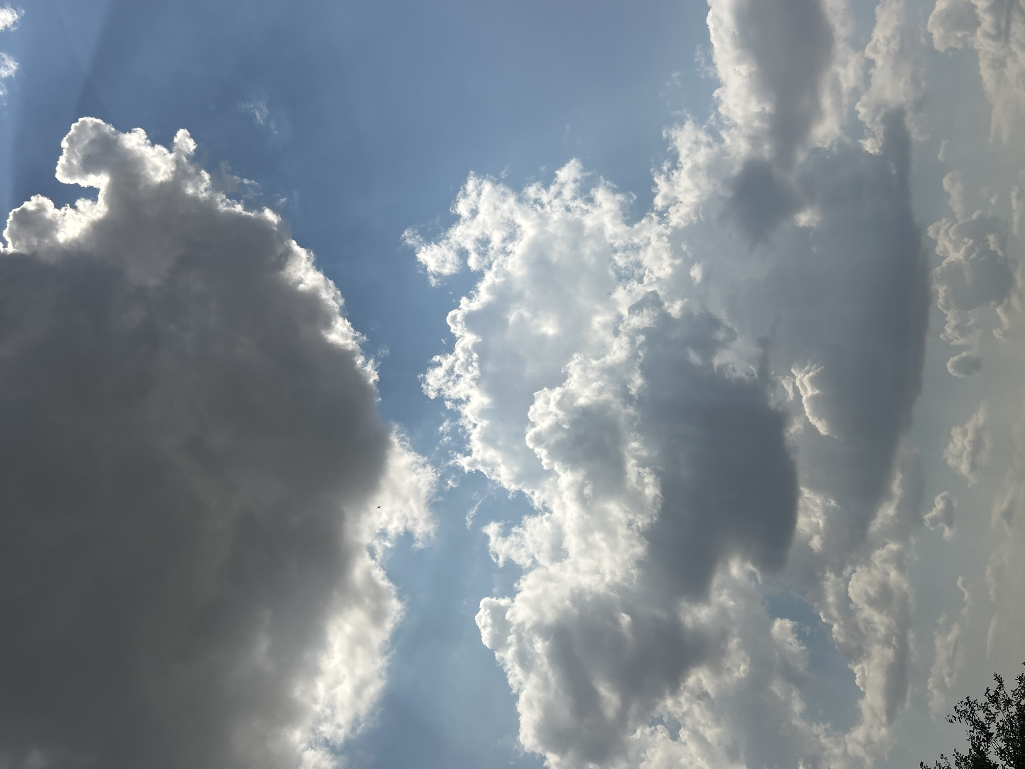 The media file shows a cloudy sky with the sun partially visible, creating dramatic light rays. The clouds vary in density, with some appearing dark and heavy, suggesting potential rain, while others are bright white and puffy, illuminated by the sun. The sky is a clear blue, peeking through the clouds in patches. The overall scene evokes a sense of grandeur and the dynamic nature of weather. The lighting suggests it is daytime, likely late afternoon given the angle of the sun's rays. There are no visible people or objects, and no text. The image appears to be a photograph taken from the ground looking upwards into the sky. The provided location context of Загорци, Bulgaria, indicates a rural or semi-rural setting.