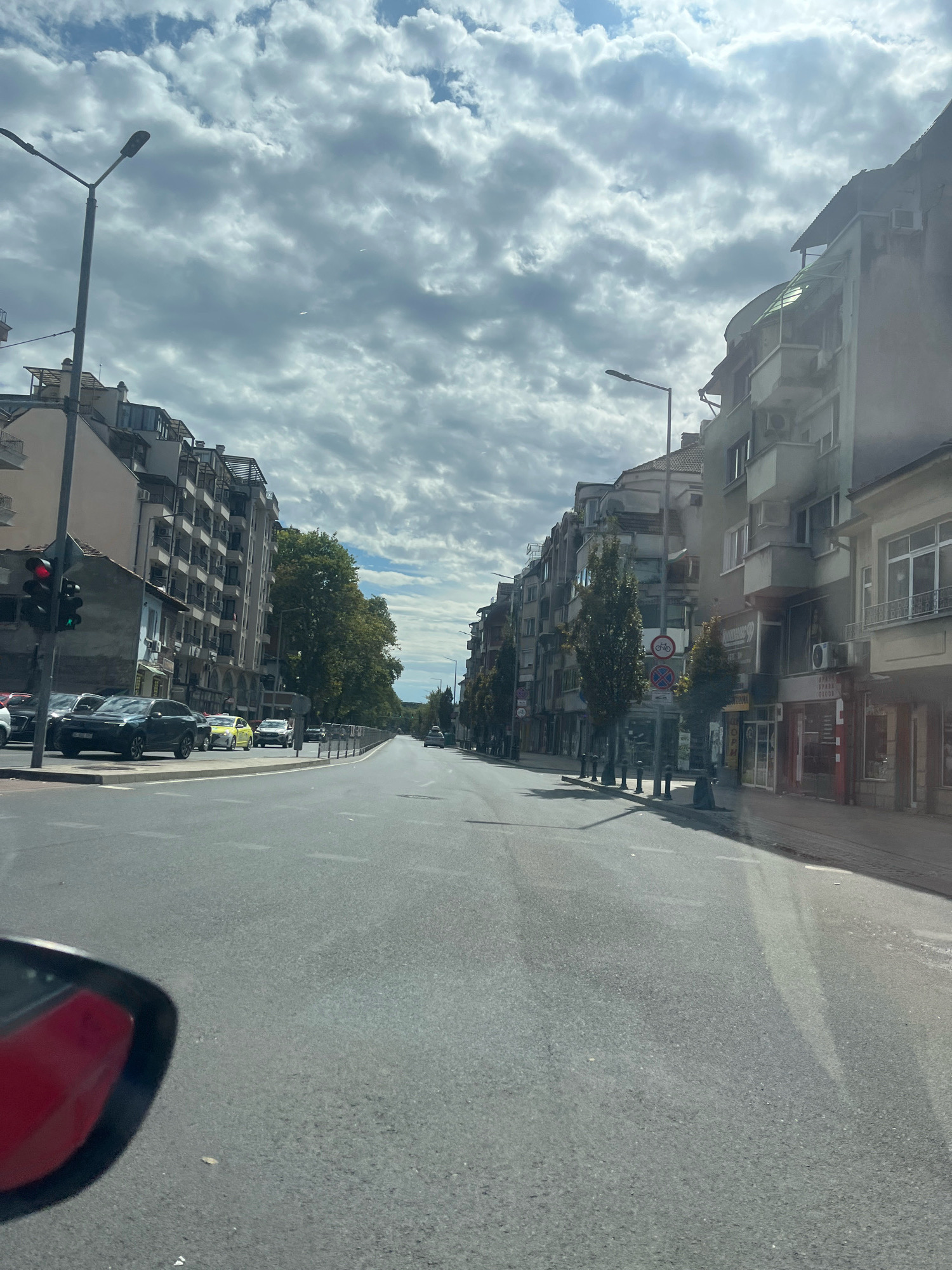 The image shows a street scene in Plovdiv, Bulgaria. The perspective is from inside a vehicle, looking forward. The street is paved and appears to be a main thoroughfare with multiple lanes. On the left side of the street, there are multi-story buildings with balconies and apartments, as well as shops on the ground floor. A traffic light is visible on the left, showing a red light. Several cars are parked or driving on the street, including a dark SUV and a yellow car. Trees line the sidewalk in places. On the right side of the street, there are also multi-story buildings with shops on the ground floor. Streetlights are present on both sides of the road. The sky is partly cloudy with a bright sun, creating some shadows. The scene depicts a typical urban environment with active traffic and pedestrian areas.