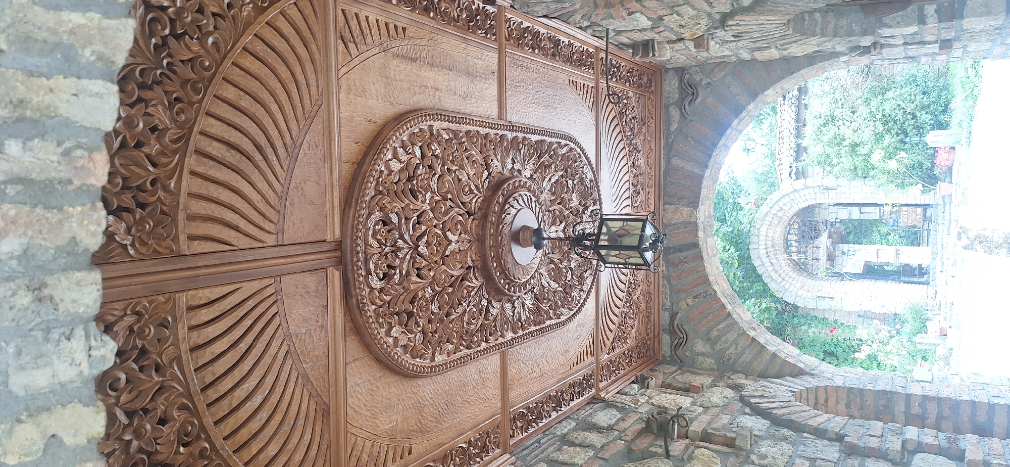 This image shows the interior of an arched entryway constructed of stone and brick. The ceiling is elaborately carved from wood, featuring floral patterns and radiating lines. A decorative lantern hangs from the center of the ceiling. Through the archway, a courtyard with greenery and more stone structures is visible. The location is identified as the Monastery of Saint George in Kalimantsi, Bulgaria. There are no people or significant actions depicted in the scene.