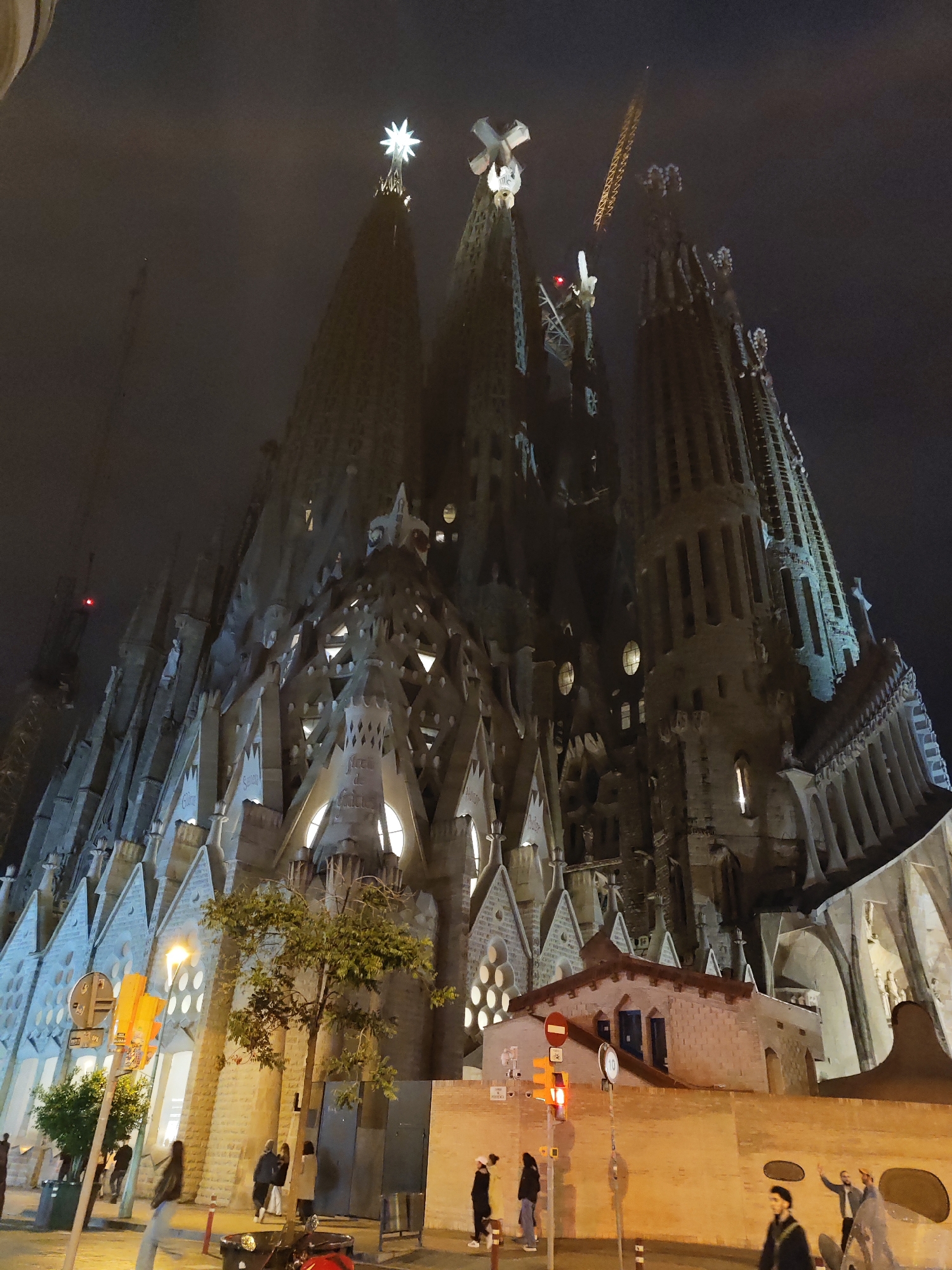 This image shows the Sagrada Familia in Barcelona, Spain, at night. The facade of the basilica is visible, including multiple towers and intricate detailing, illuminated by artificial light. A construction crane is visible towards the top of the building. At the bottom of the image are people walking on the sidewalk. Also at the bottom are street signs, a tree, a brick wall, and a small building with a red stop sign. The sky is dark, possibly indicative of fog or nighttime conditions.