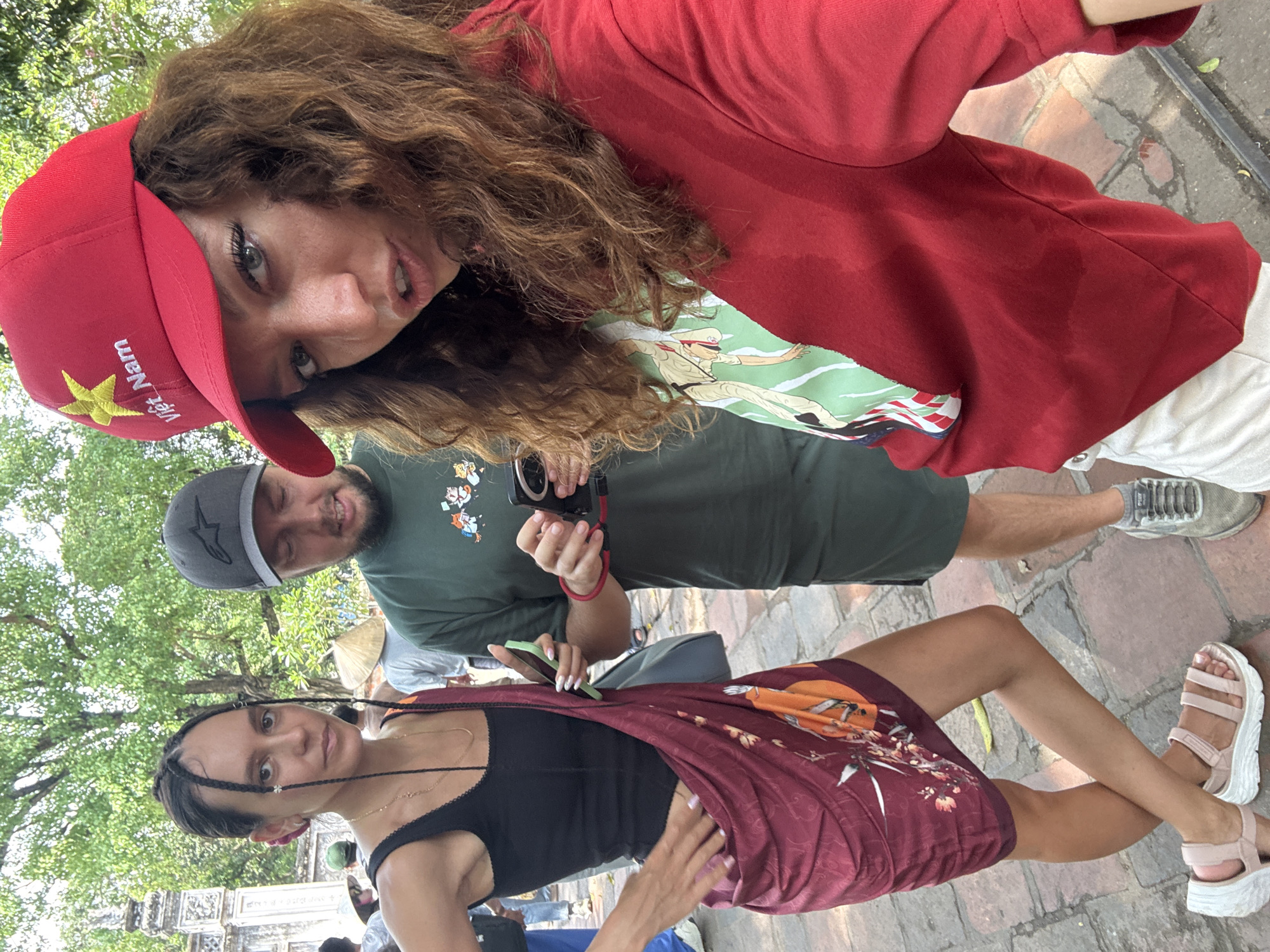 Three people are posing for a photo outdoors on a brick-paved ground. A woman with curly brown hair and a red cap labeled "Viet Nam" in yellow is on top, angled towards the camera. Below her is a man holding a camera and looking towards the woman in the lower part of the frame. The woman in the lower part has braided hair and is wearing a dark top and a patterned skirt. Trees and architecture can be seen in the background. The location may be Hà Nội, Vietnam, based on the cap's label.