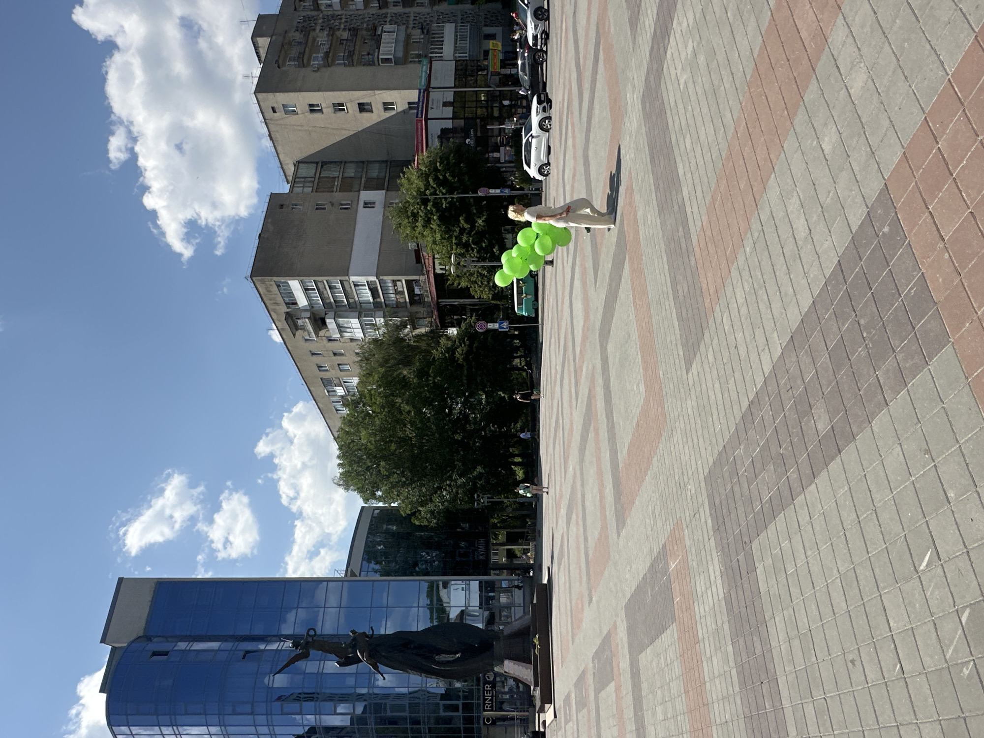 A medium shot captures a sunny day in Ternopil, Ukraine, likely on Teatralna Square. In the foreground, a person in a light-colored outfit walks from right to left, holding a bunch of green balloons. The ground is paved with a patterned arrangement of grey and reddish-brown rectangular stones. In the midground, several cars are parked and driving, including a white SUV. To the left, a tall, dark sculpture of figures, possibly historical or mythological, stands before a modern building with a blue, glass facade. Behind this, trees and several apartment buildings with balconies are visible under a bright blue sky with scattered white clouds. A turquoise van is also visible in the background to the left.