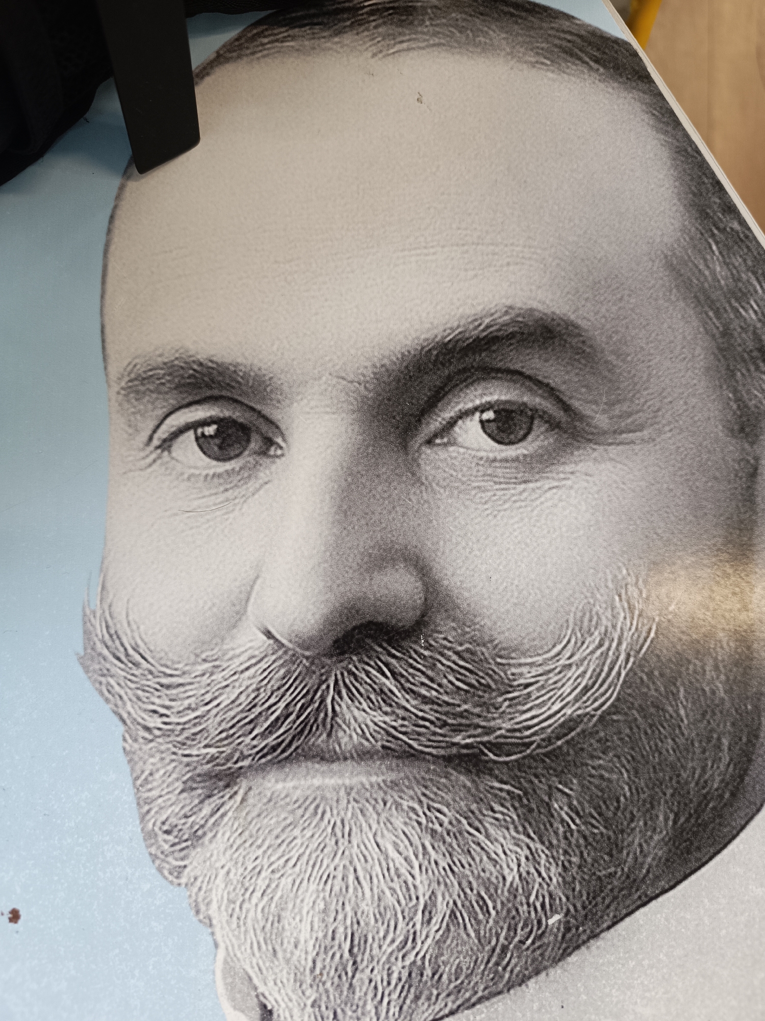 A close-up, black-and-white image depicts a man with a prominent mustache and beard. The image appears to be a portrait or a reproduction of a portrait, possibly on paper. Details of the face, including the eyes, nose, mustache, and beard, are visible. The background is light blue. Part of a black object appears at the top left of the image. The photograph was taken in Pleven, Bulgaria.