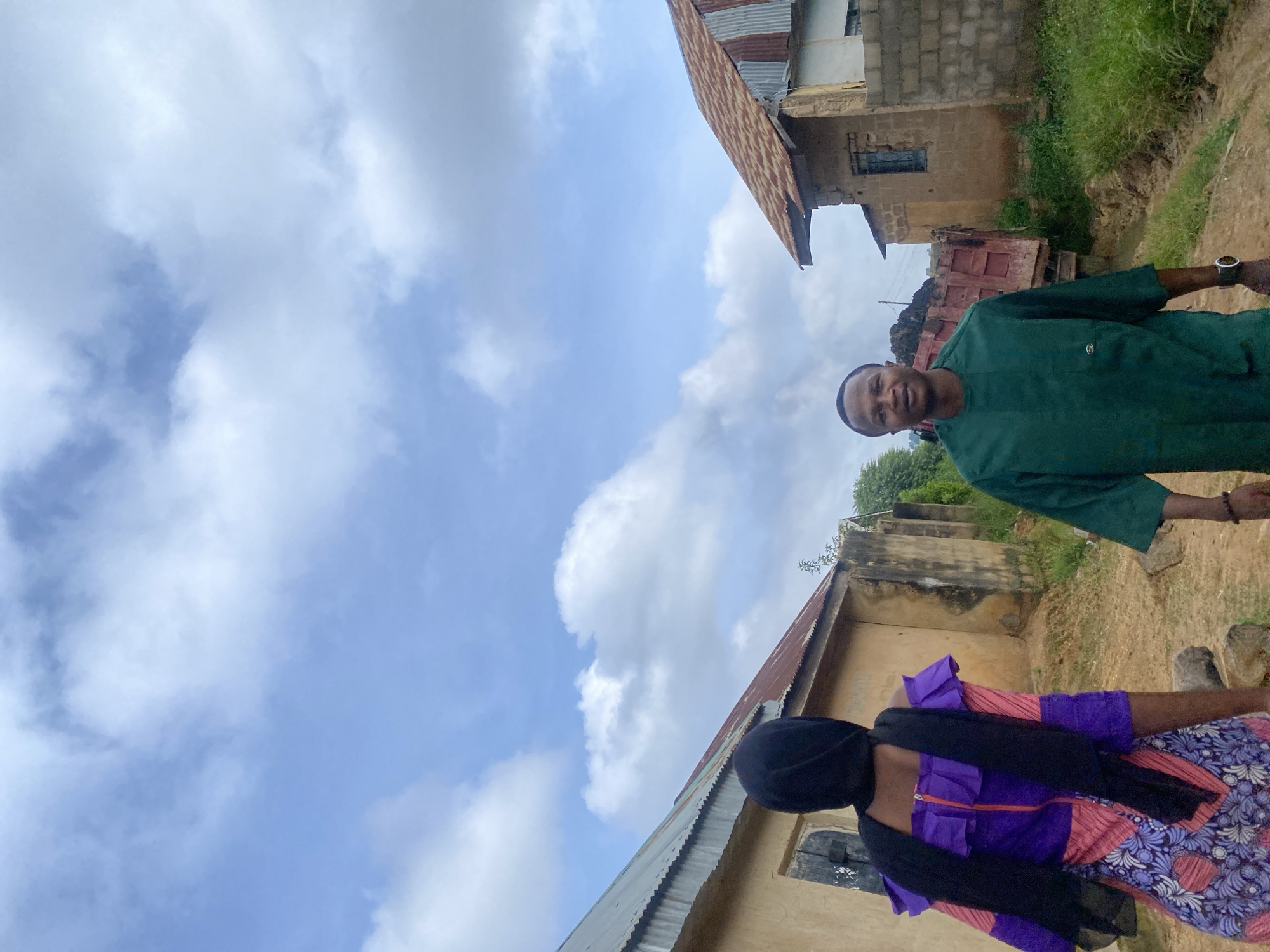 The image depicts an outdoor scene in Rafin Par, Nigeria, under a partly cloudy sky. Two individuals are visible. In the foreground, a person is walking away from the camera, wearing a purple and pink patterned dress with a black headscarf and a black scarf draped around their neck and shoulders. To the right, a man is facing the camera, smiling and wearing a green tunic. Behind him, a red dump truck is partially visible, loaded with dark material, suggesting construction or hauling activity. The background features simple buildings with corrugated metal roofs and rough plaster walls, typical of rural or developing urban environments. The ground appears to be dirt or unpaved road.