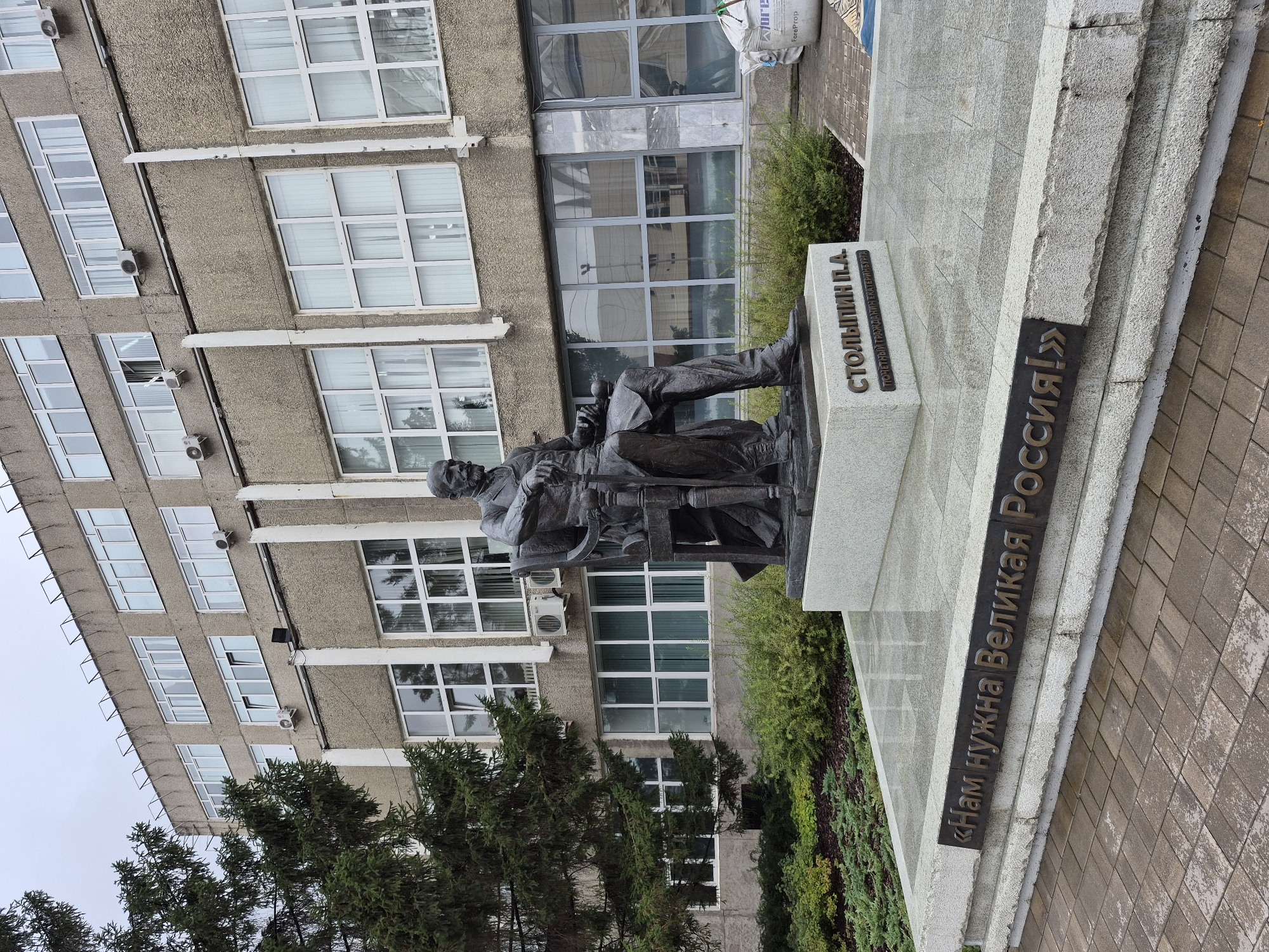 The image shows a bronze statue of Pyotr Stolypin seated on a chair, located in Yekaterinburg, Russia. The statue is placed on a granite pedestal inscribed with "СТОЛЫПИН П.А." (Stolypin P.A.) and "ПОЧЕТНЫЙ ГРАЖДАНИН ЕКАТЕРИНБУРГА" (Honorary Citizen of Yekaterinburg). Below this, a dark metal plaque bears the inscription in Russian: "«Нам нужна Великая Россия!»" ("We need a Great Russia!"). The statue is situated in front of a brutalist concrete building with rows of windows. To the left of the statue is a large evergreen tree, and in front of the building, there is a landscaped area with bushes and some bags of construction material. The ground in front of the monument is paved with brick tiles. The sky is overcast, suggesting a cloudy day.