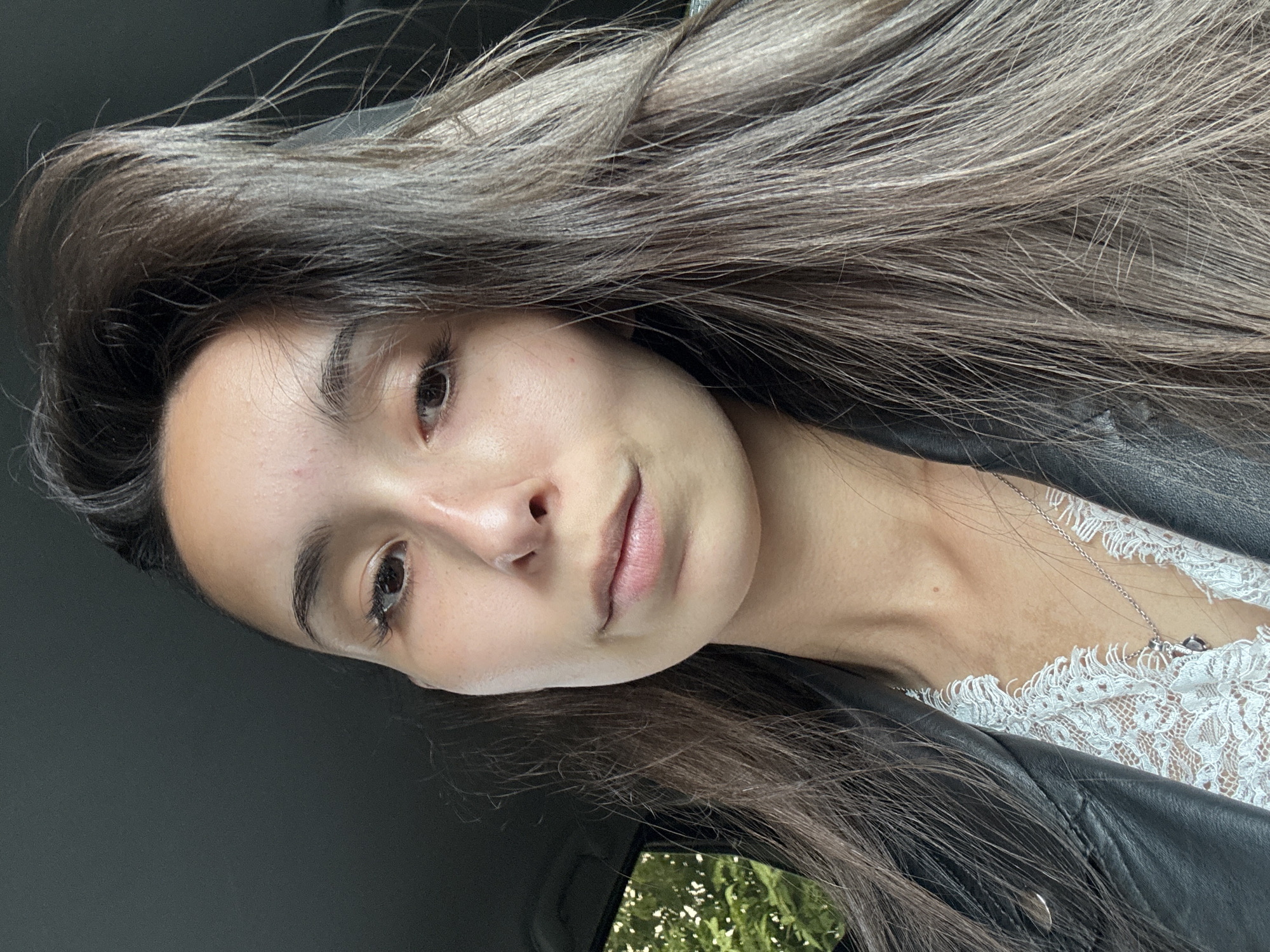 A close-up selfie, taken from a low angle, shows a young woman with long, dark brown hair styled with volume at the crown. She is wearing a white lace top beneath a black leather jacket. A delicate silver necklace with a small pendant is visible. The background is dark, with a hint of greenery visible through a window on the left side, suggesting the car's interior. The overall lighting is soft, illuminating her face, which shows natural makeup with a focus on her eyes and lips. No specific landmarks or indicators of the city of Izhevsk, Russia, are present in the image.