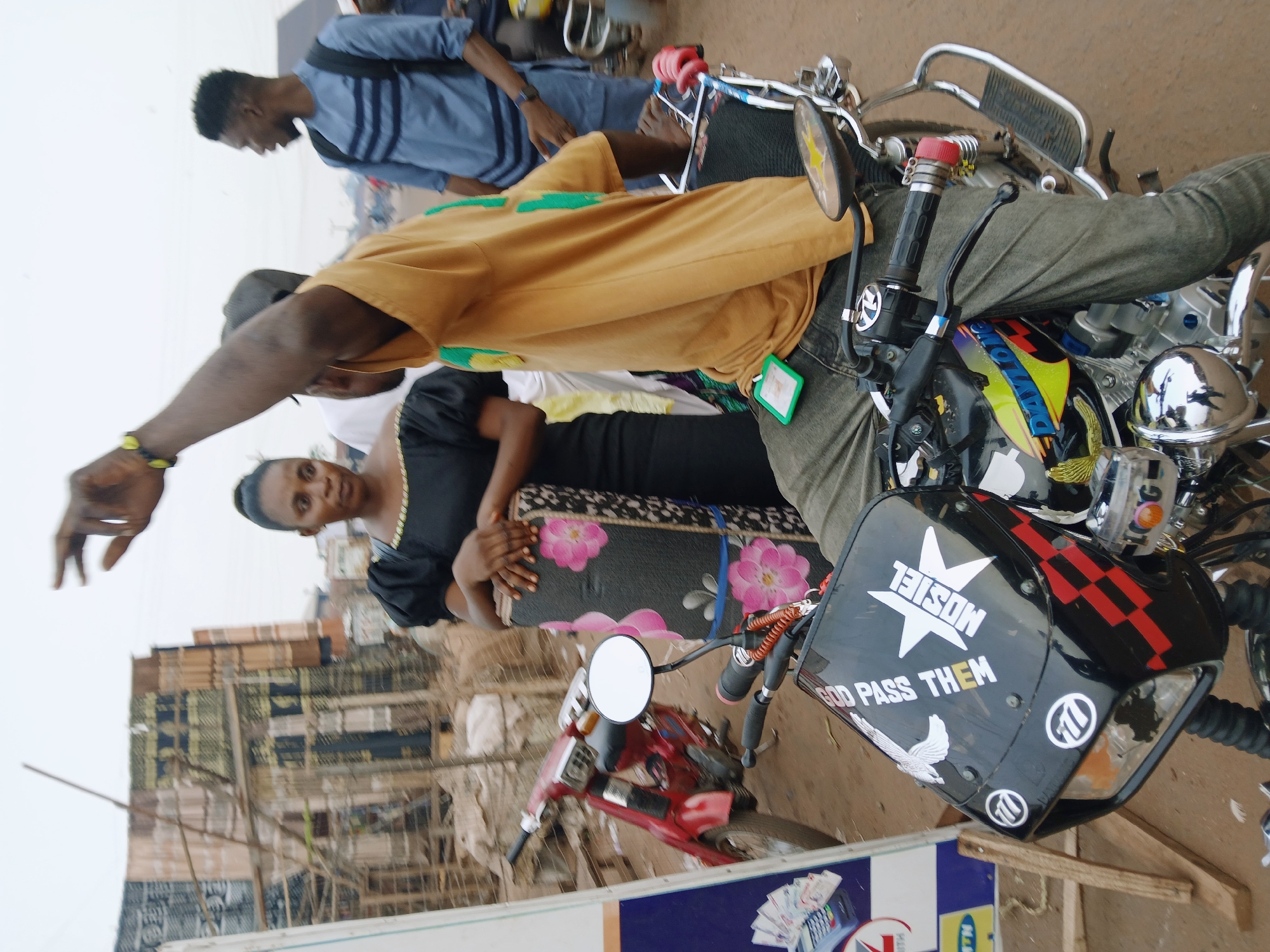The image shows a scene in Ankpa, Nigeria. A woman in a black dress stands next to a motorcycle, holding a patterned bag with floral designs. A man in a yellow shirt is seen standing next to her, his arm raised. Another man in a blue shirt is visible in the background. In the foreground, there's another motorcycle with a custom fairing, displaying a "Mosiel" logo and the text "GOD PASS THEM." Buildings are visible in the background.