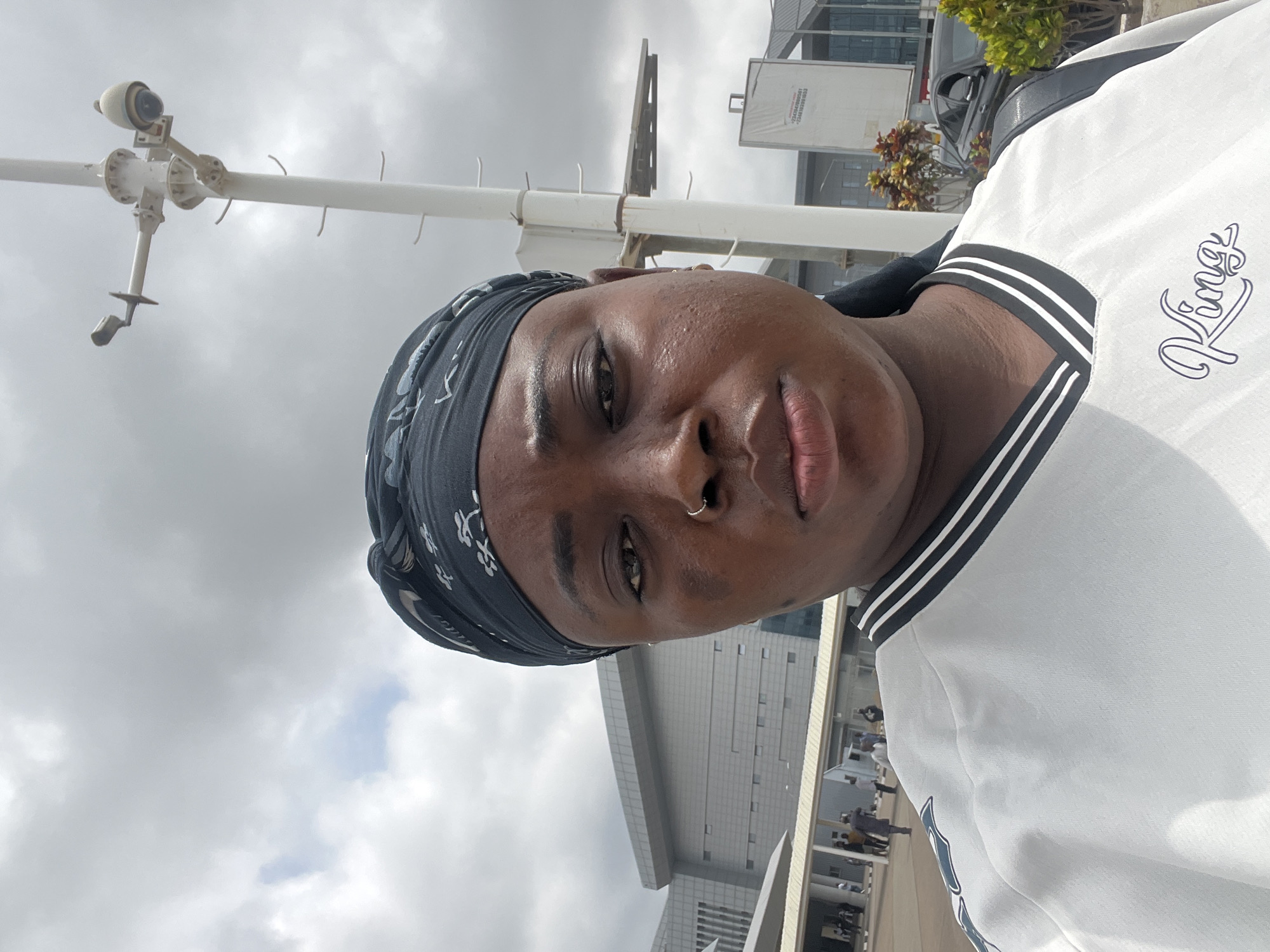A close-up, waist-up portrait captures an individual in an outdoor setting, taken from a low angle and rotated 90 degrees clockwise. The subject is a person of color, looking directly into the camera with a neutral expression. They wear a dark, patterned bandana covering their head and a white top featuring black and white stripes around the collar. The word "Kings" is visibly embroidered in a stylized font on the left side of their chest (viewer's right). A small silver nose ring adorns their right nostril.
The background is dominated by an overcast sky filled with grey clouds. A prominent white pole, fitted with a surveillance camera or weather monitoring equipment, stands to the left of the subject. This equipment includes a spherical camera component and several arm-like extensions. In the mid-ground, modern buildings with glass exteriors are visible, suggesting an urban or commercial area. A dark vehicle and some green foliage are partially seen further back on the right. Distant, blurry figures can be discerned near one of the buildings. The photograph depicts a daytime scene under cloudy weather conditions in Gwaska, Nigeria.
