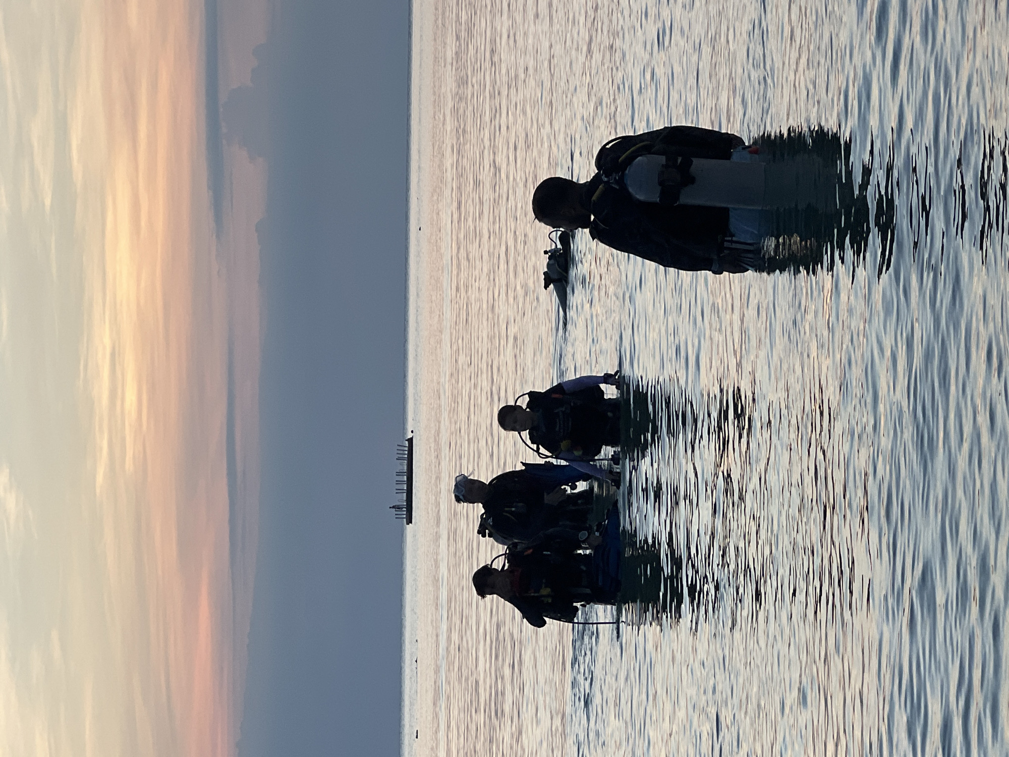 This image depicts a group of scuba divers in shallow, calm water during what appears to be sunset. The sky displays hues of pink, orange, and blue, indicative of twilight. In the background, a long, flat barge with vertical structures, possibly pilings or poles, is visible on the horizon. In the foreground, five individuals, all wearing scuba gear including tanks and BCDs, are wading through the water. One diver is closest to the camera, facing away, and is silhouetted against the water. To their left and further back, three other divers are grouped together, facing the camera. Another diver is partially submerged and swimming further out. The water is clear, and the reflections of the divers and the sky are visible on its surface. The location is identified as Ko Pha-ngan District, Thailand, a region known for its tropical waters and diving opportunities.