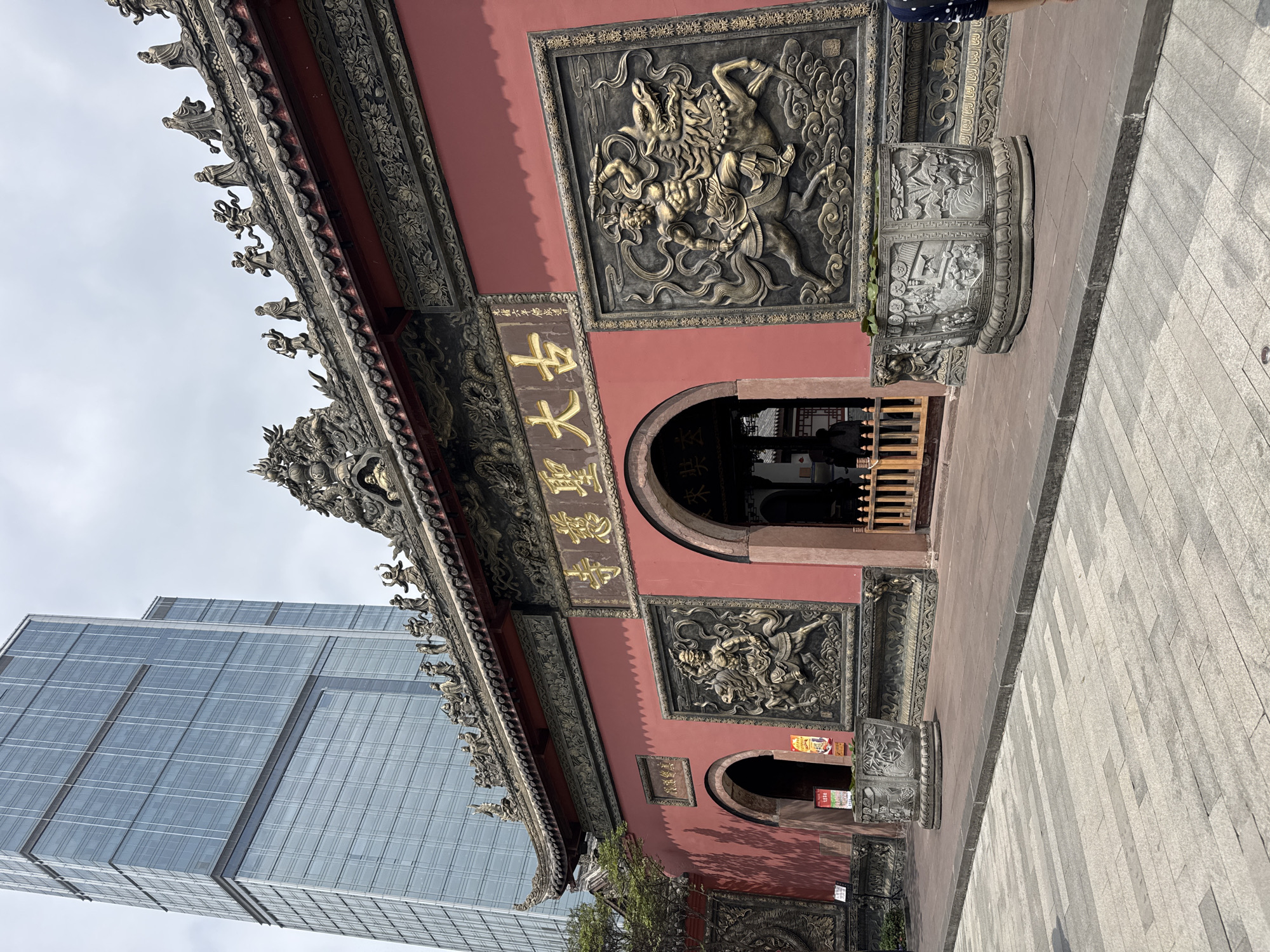 The image shows the main building of the Chengdu Daci Temple in Jinjiang District, China. The building has a traditional Chinese architectural style with a red exterior, ornate carvings, and a tiled roof. A large modern skyscraper can be seen behind the temple, contrasting the old and new structures. The carvings on the temple depict mythical creatures and figures, and golden Chinese characters are visible above the entrance. Two stone planters are positioned on either side of the entrance, adorned with intricate designs. The ground in front of the temple is paved with grey stone tiles.