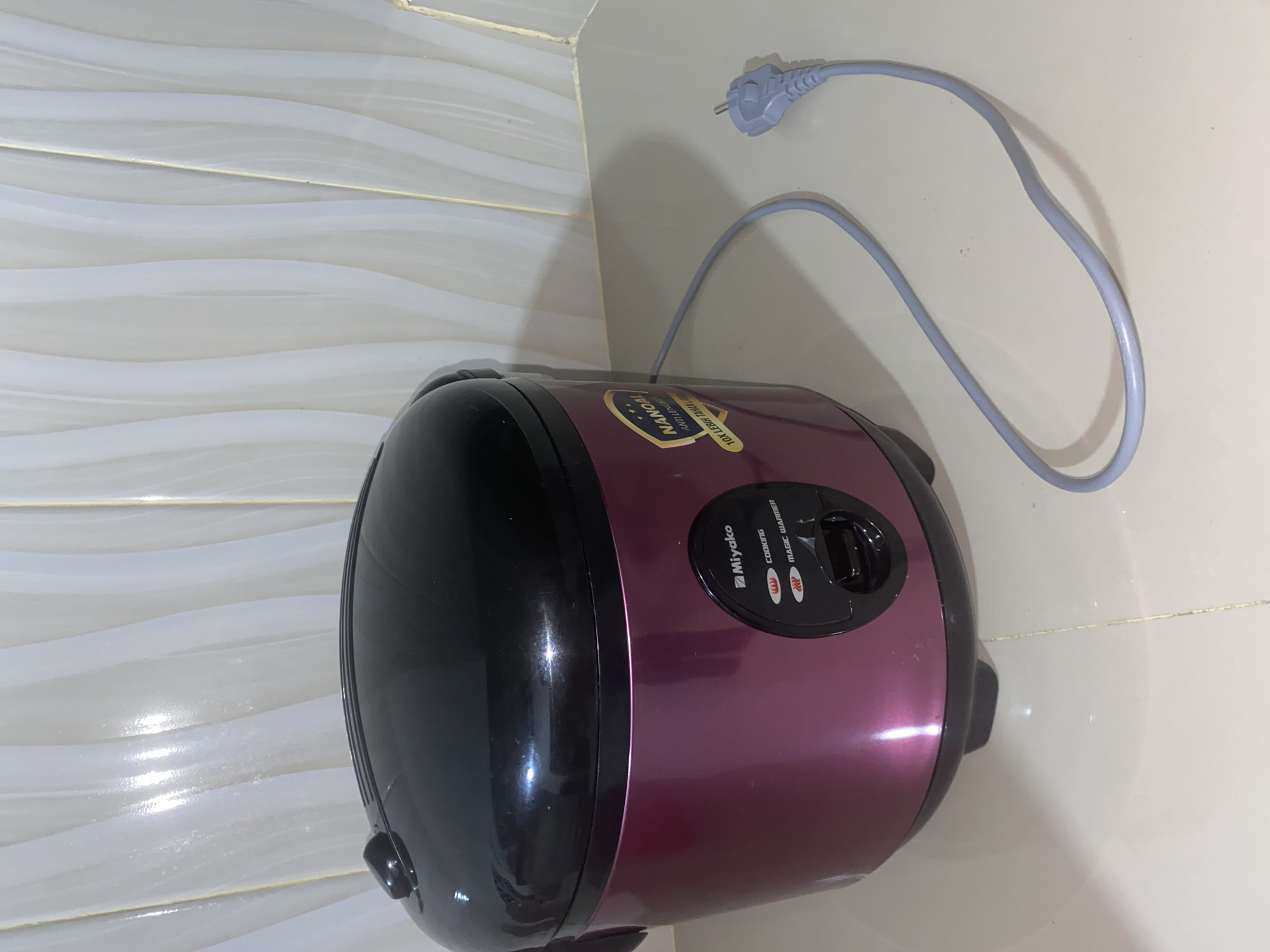 A purple and black rice cooker sits on a tiled countertop in Bontoharu, Indonesia. The rice cooker has a black lid and a purple body with a control panel featuring "Miyako," "Cooking," and "Magic Warmer" labels. A grey electrical cord extends from the rice cooker to a grey electrical plug lying on the countertop. The background consists of white, wavy tiles.
