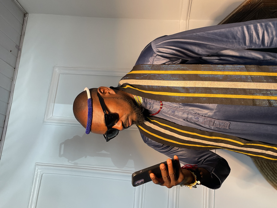 A medium shot depicts an adult male, bald with dark skin and a neatly trimmed beard, standing indoors against a light-colored paneled door in Barakin Akawo, Nigeria. He wears a dark blue long-sleeved garment, an elaborate striped scarf with black, gold, and silver vertical stripes draped over his shoulders, a blue and white beaded headband, dark rectangular sunglasses, a red beaded necklace, and multiple bracelets on his left wrist. The individual holds a black smartphone in his left hand, looking down at the screen. Strong directional light illuminates the scene from the right, casting a distinct shadow of the man onto the door behind him.